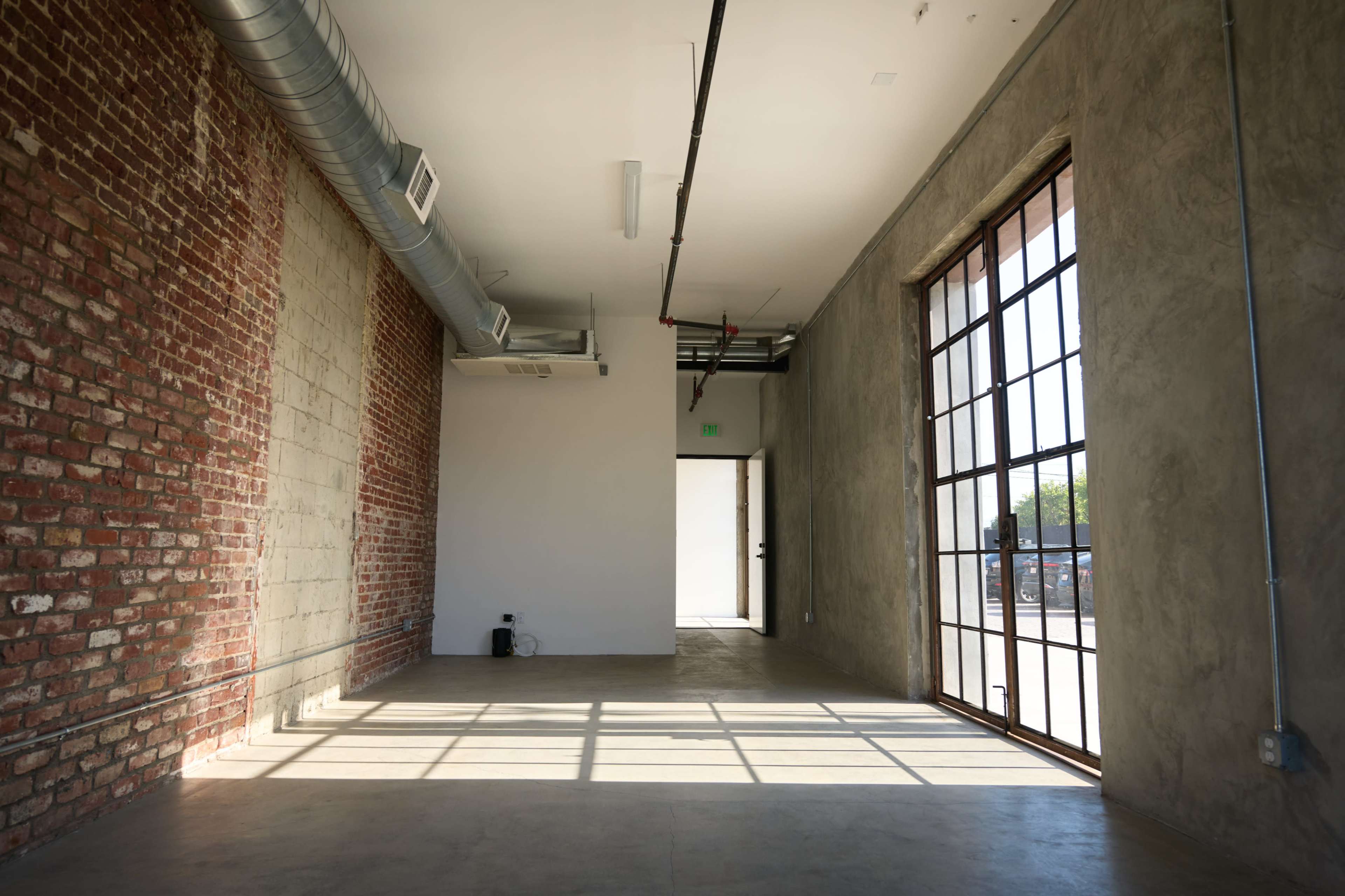The image shows an empty room with exposed brick walls and large windows allowing natural light to enter.