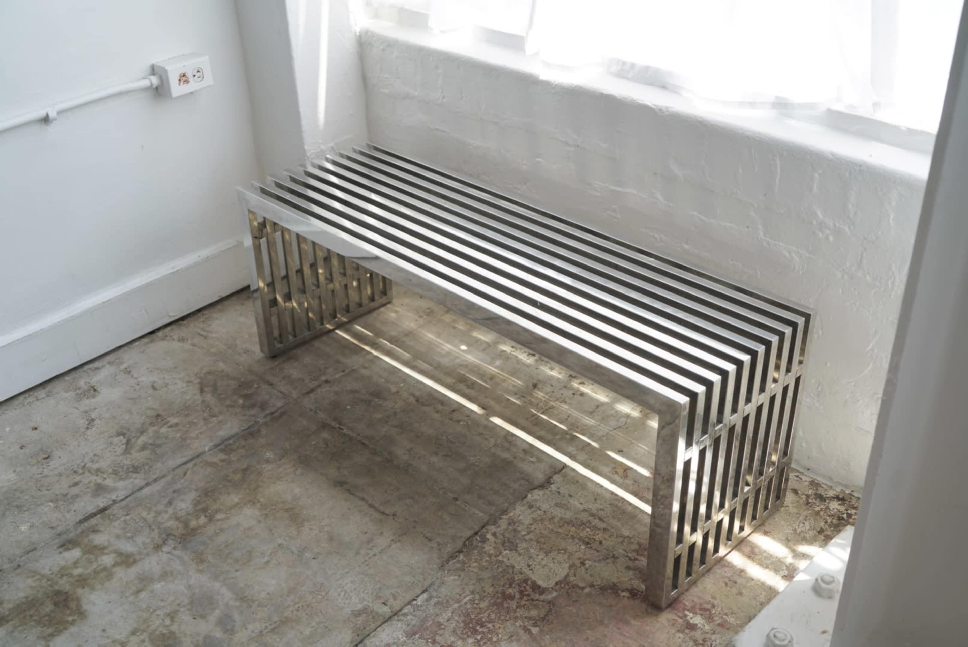 A metallic bench with slatted design is positioned beside a window on a concrete floor.