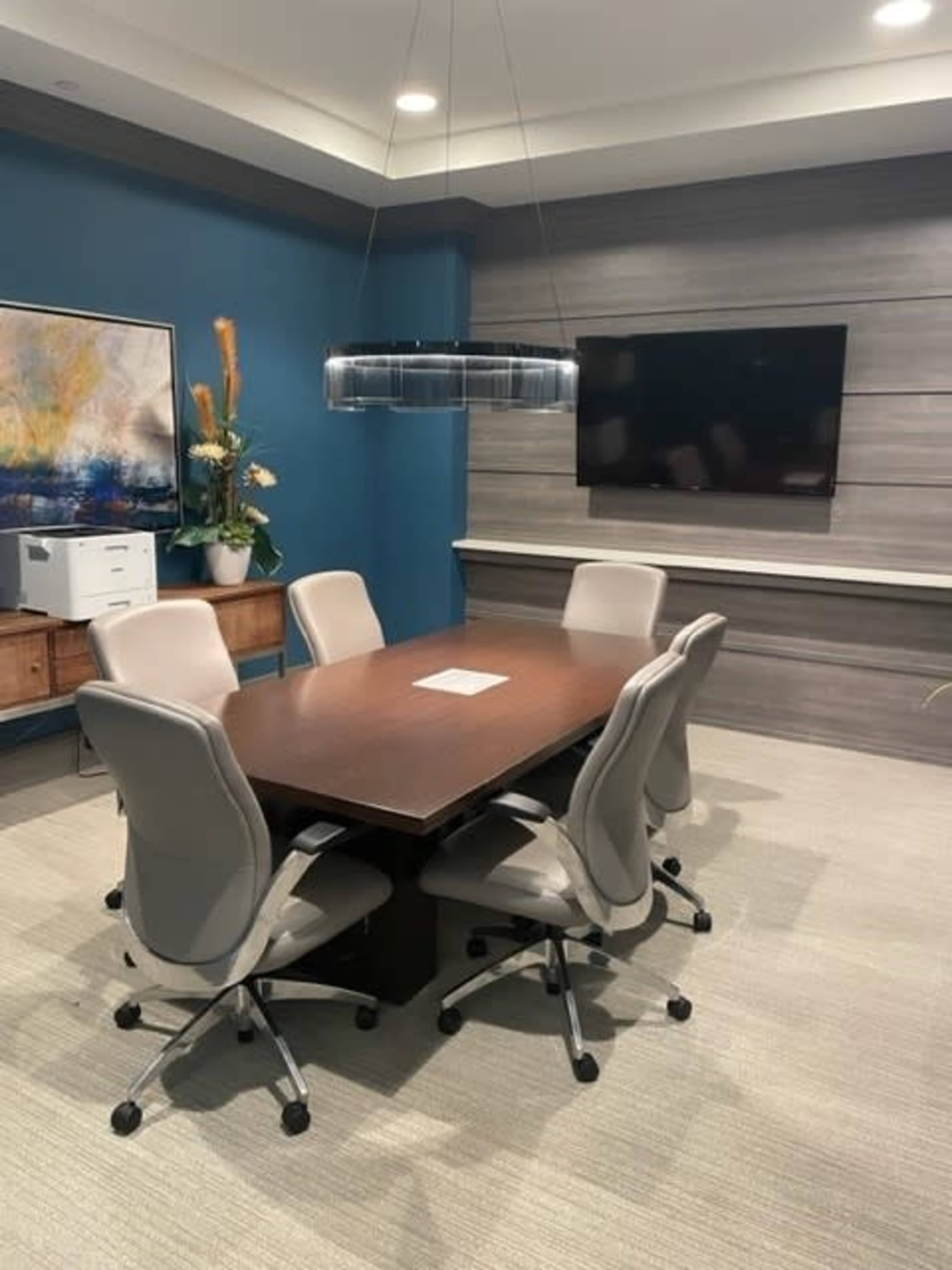 A conference room with a large wooden table surrounded by six light gray chairs, featuring a wall-mounted television and artwork.