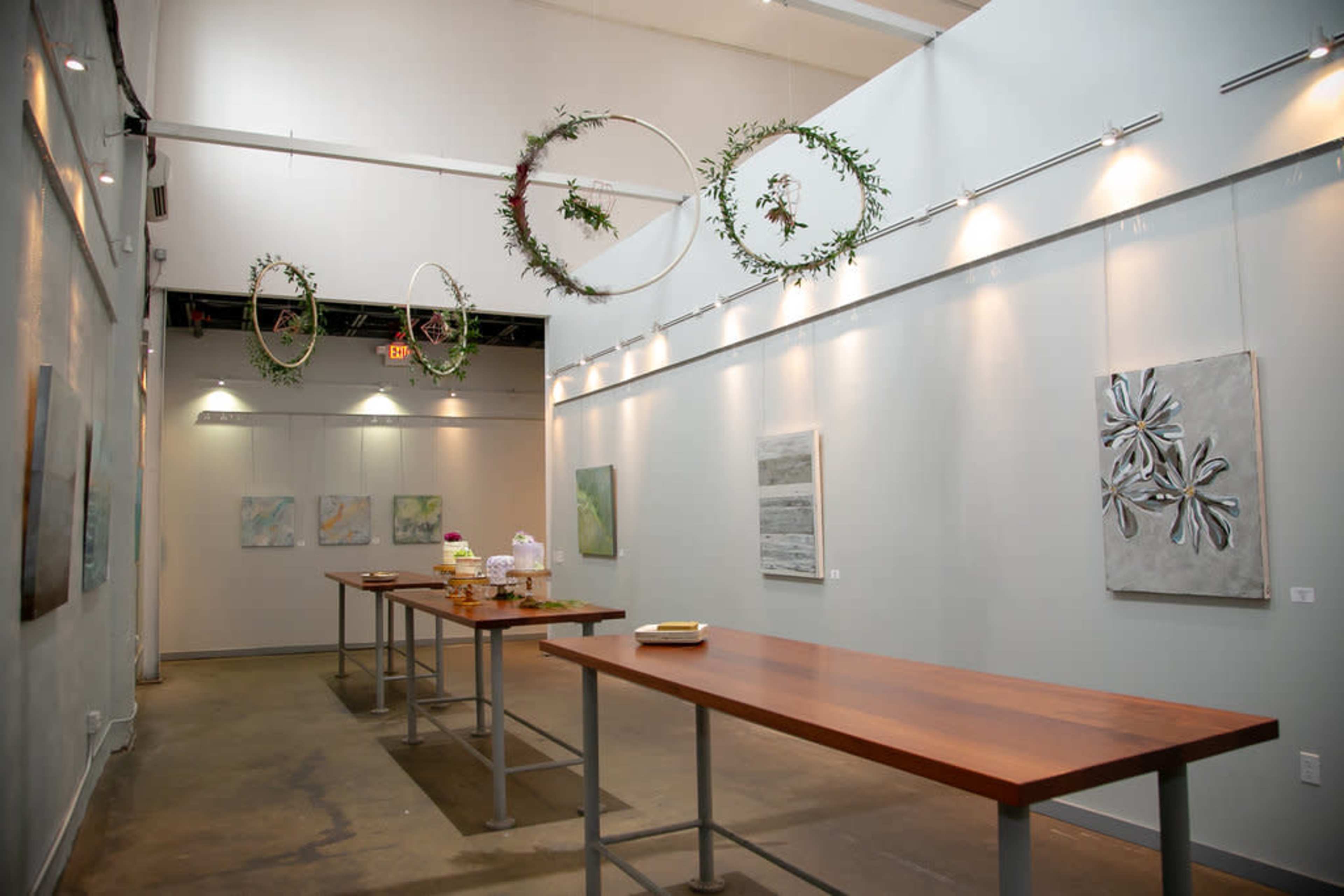 An art gallery with wooden tables set up in a spacious, well-lit room featuring hanging circular decorations and several paintings on the walls.
