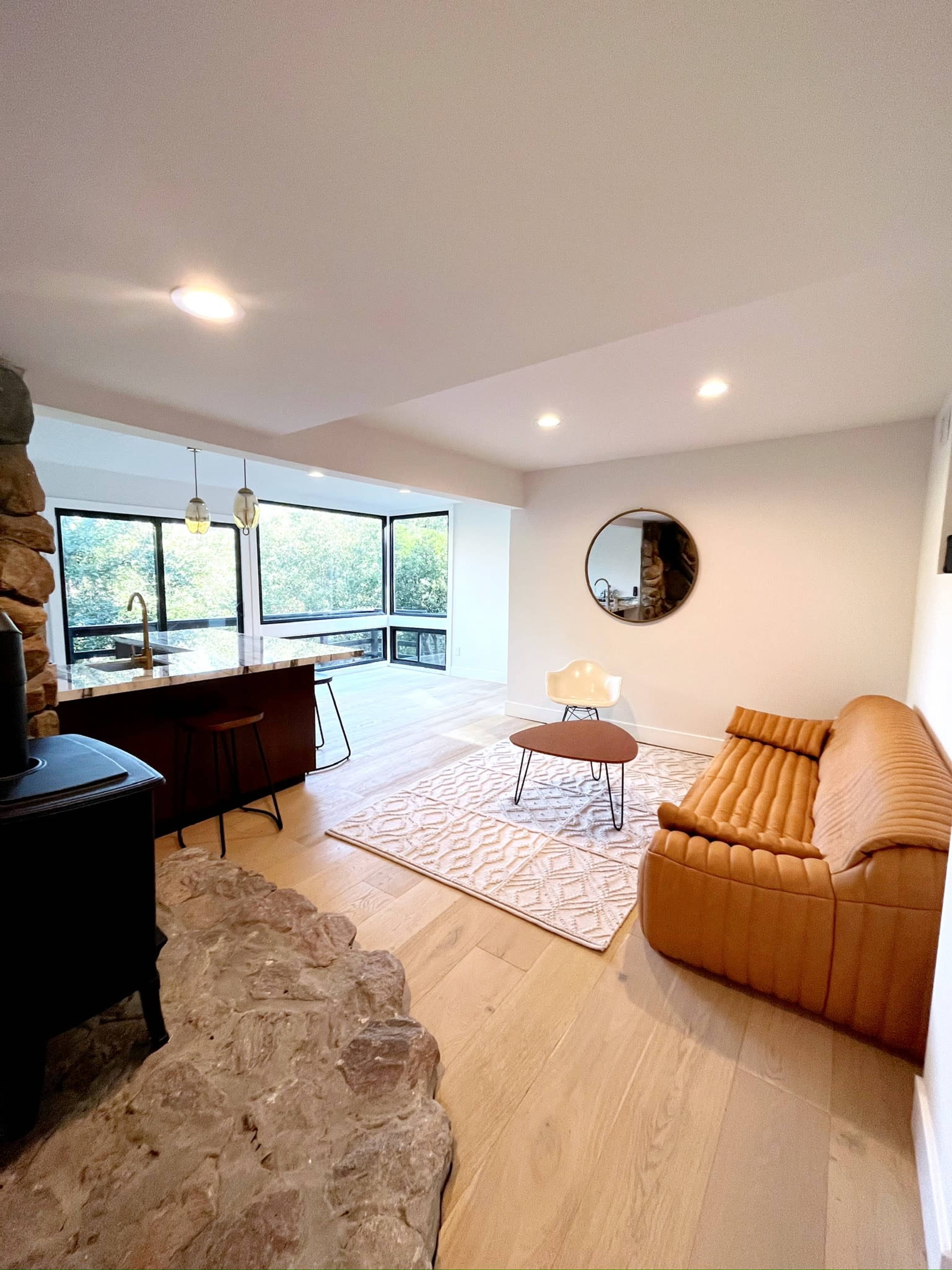 A modern living space featuring a brown couch, a round mirror on the wall, and large windows that overlook a natural landscape.