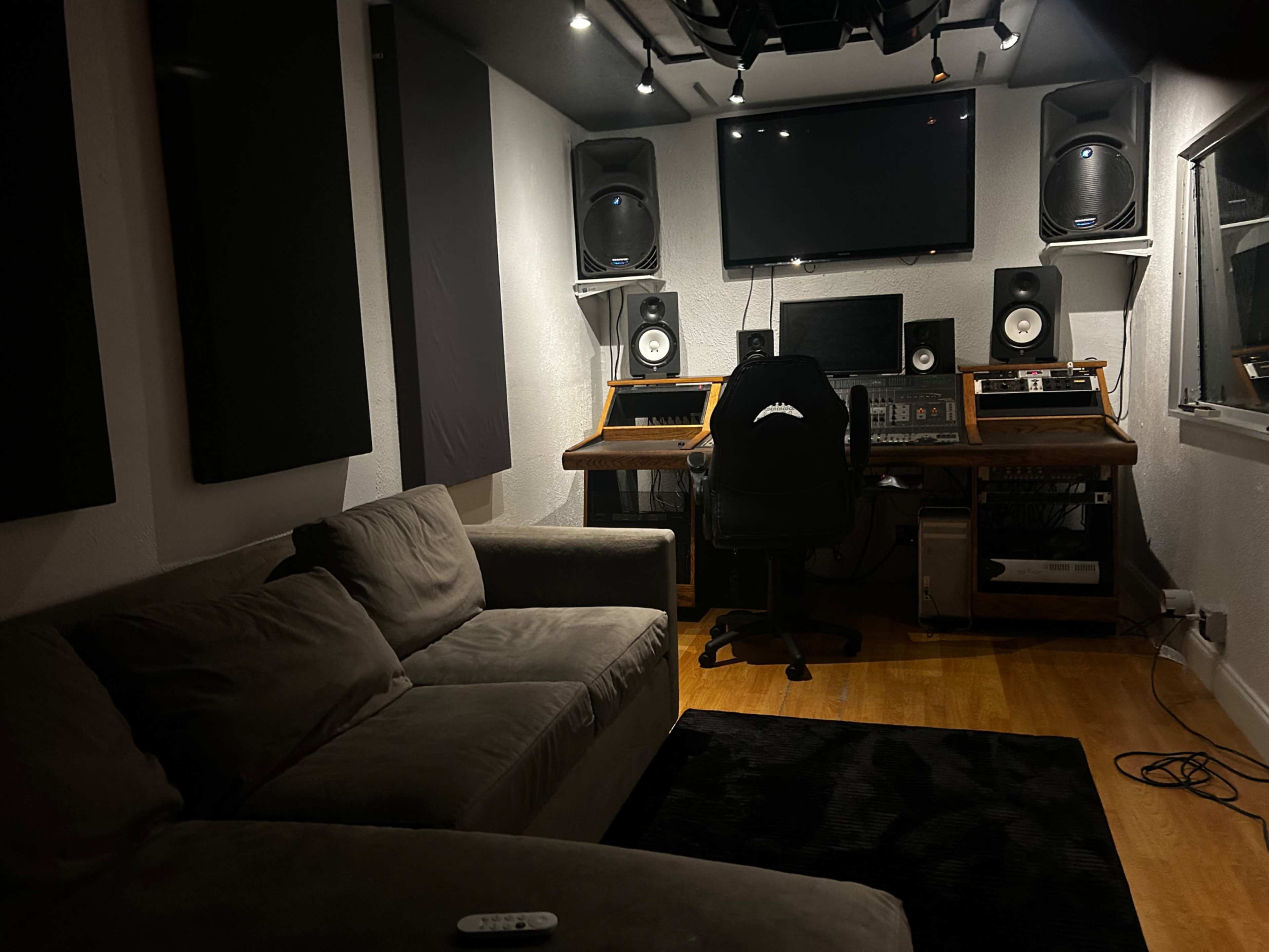 A recording studio featuring a console desk, large speakers, a computer setup, a sofa, and acoustic panels on the walls.