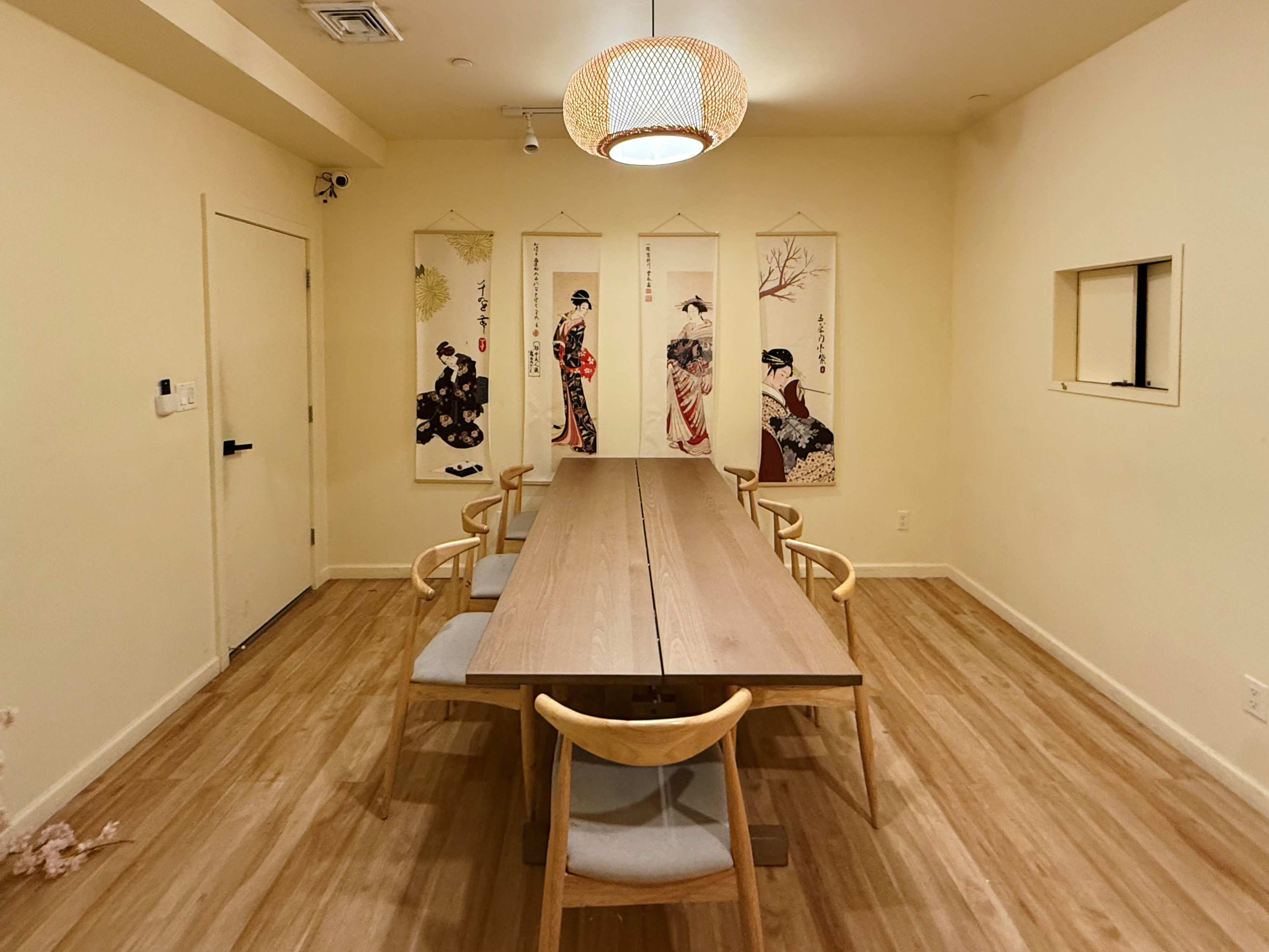 A long wooden table with six chairs is positioned in a softly lit room adorned with hanging scrolls featuring traditional Japanese images.