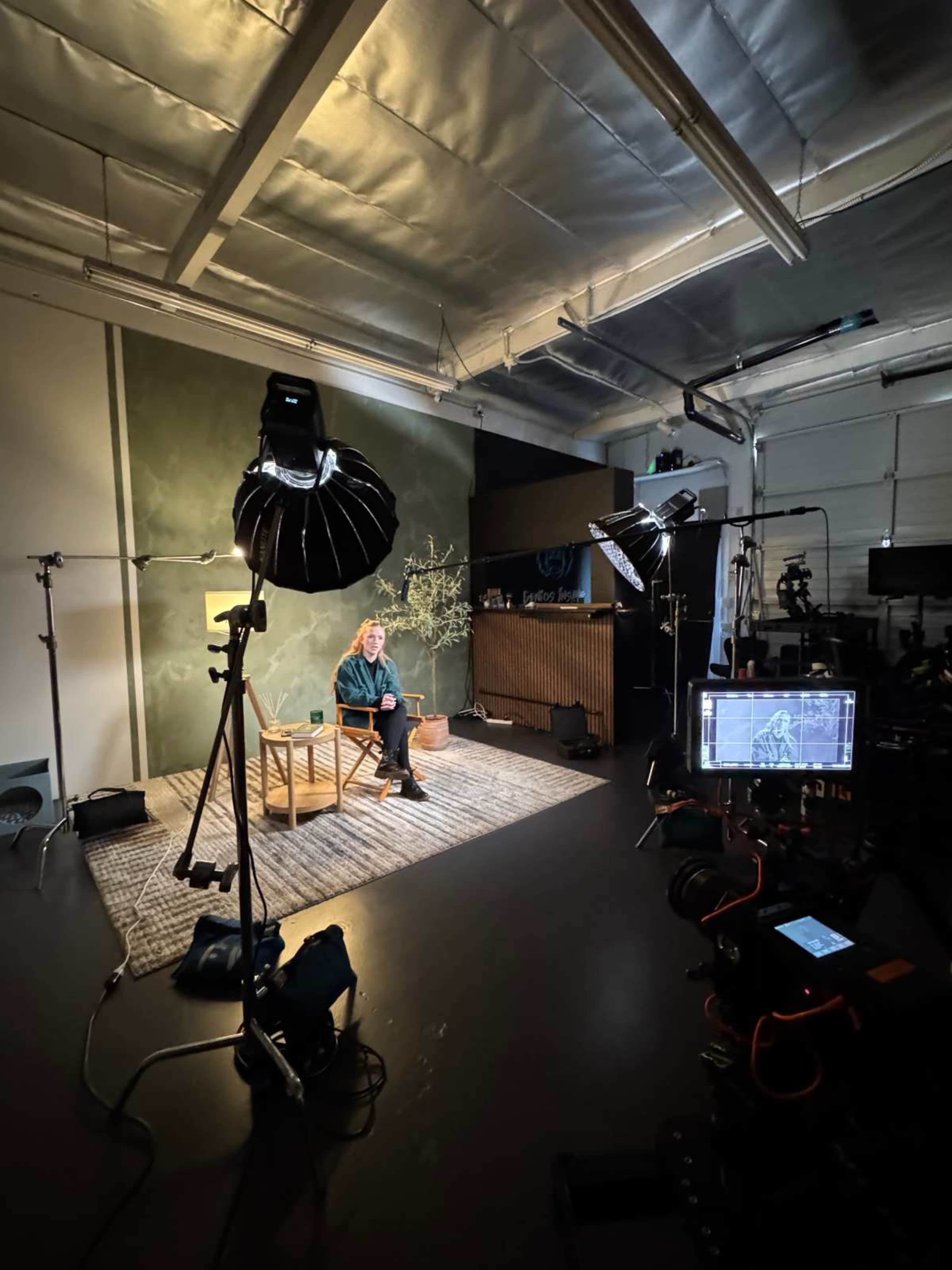 The image shows a studio setup with a person seated in a chair surrounded by lighting equipment and cameras.