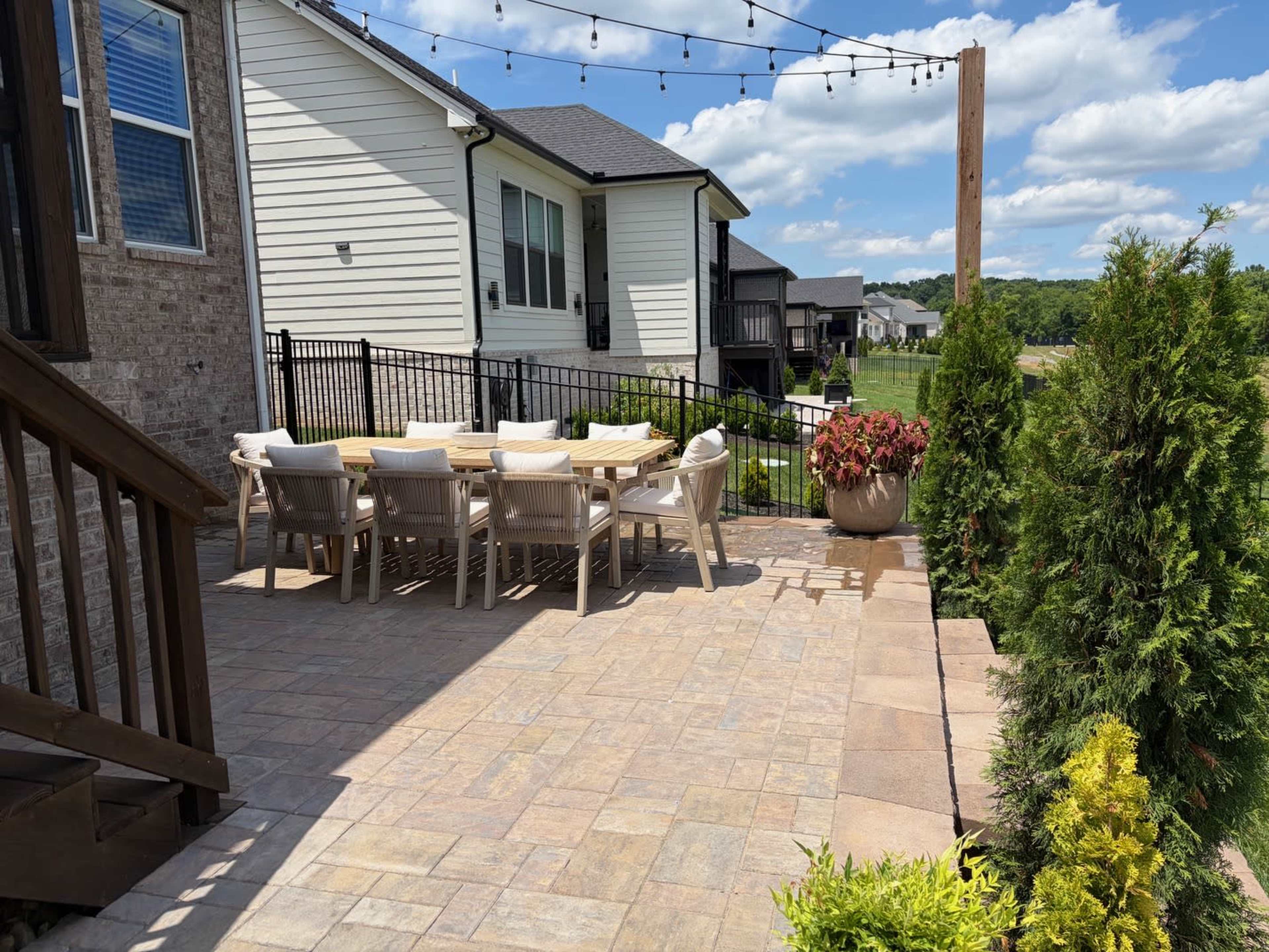 The image shows a backyard patio area with a large dining table surrounded by chairs, bordered by greenery and gently sloping terrain.