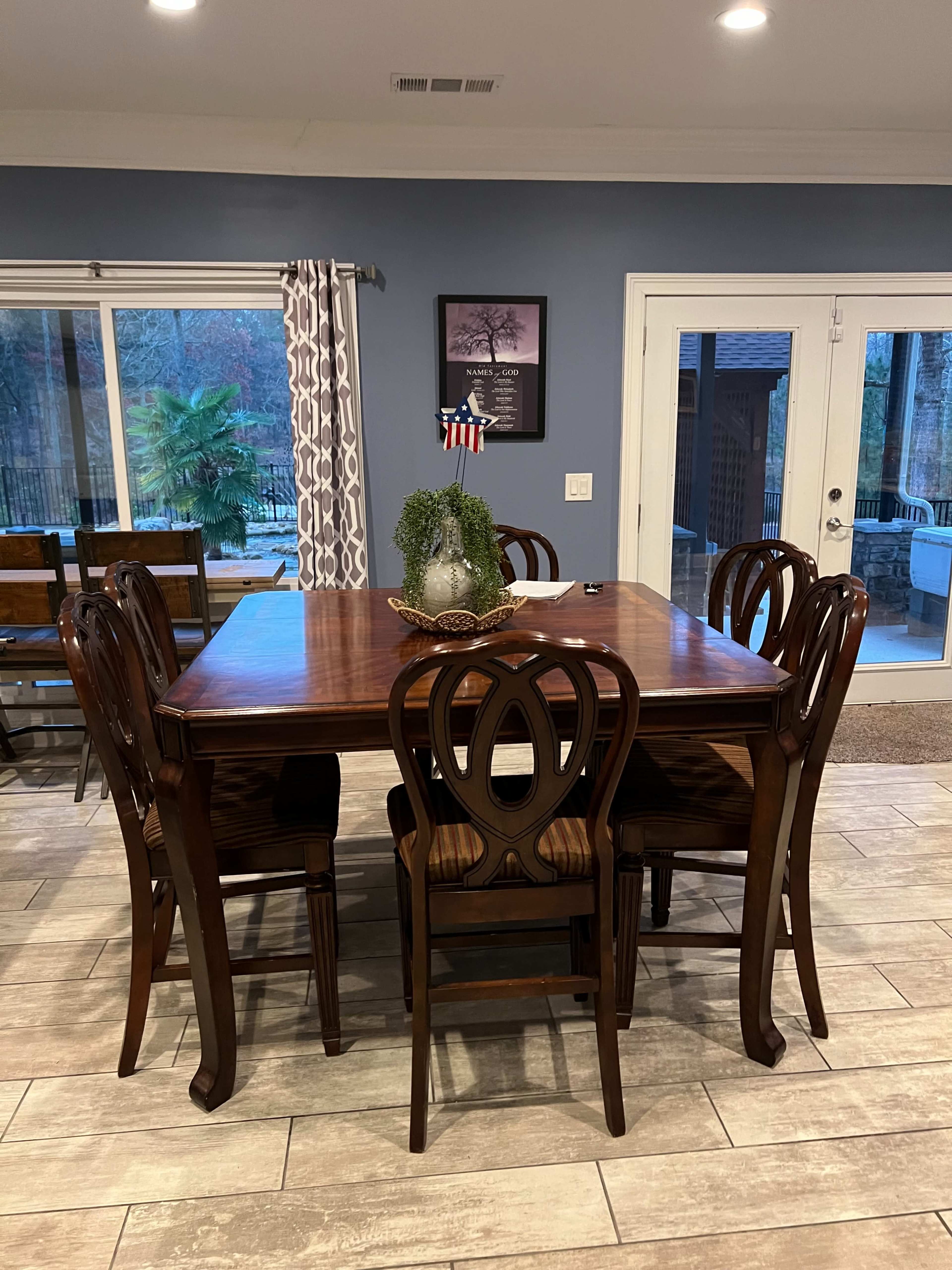 A wooden dining table with six chairs is set in a brightly lit room featuring large windows and a door that opens to a patio.