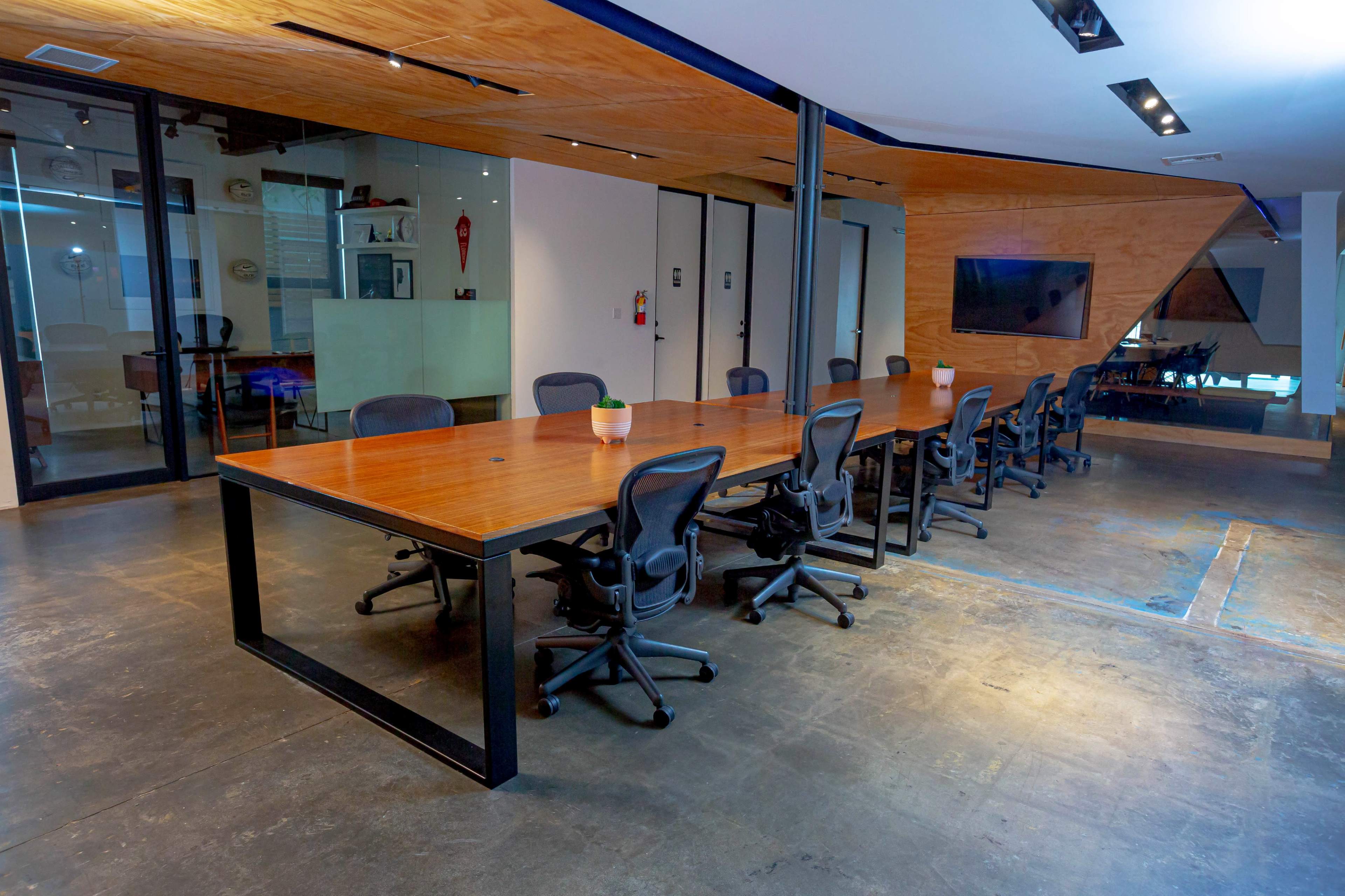 A long wooden conference table with black chairs is set in a modern meeting room with glass walls and a large screen.