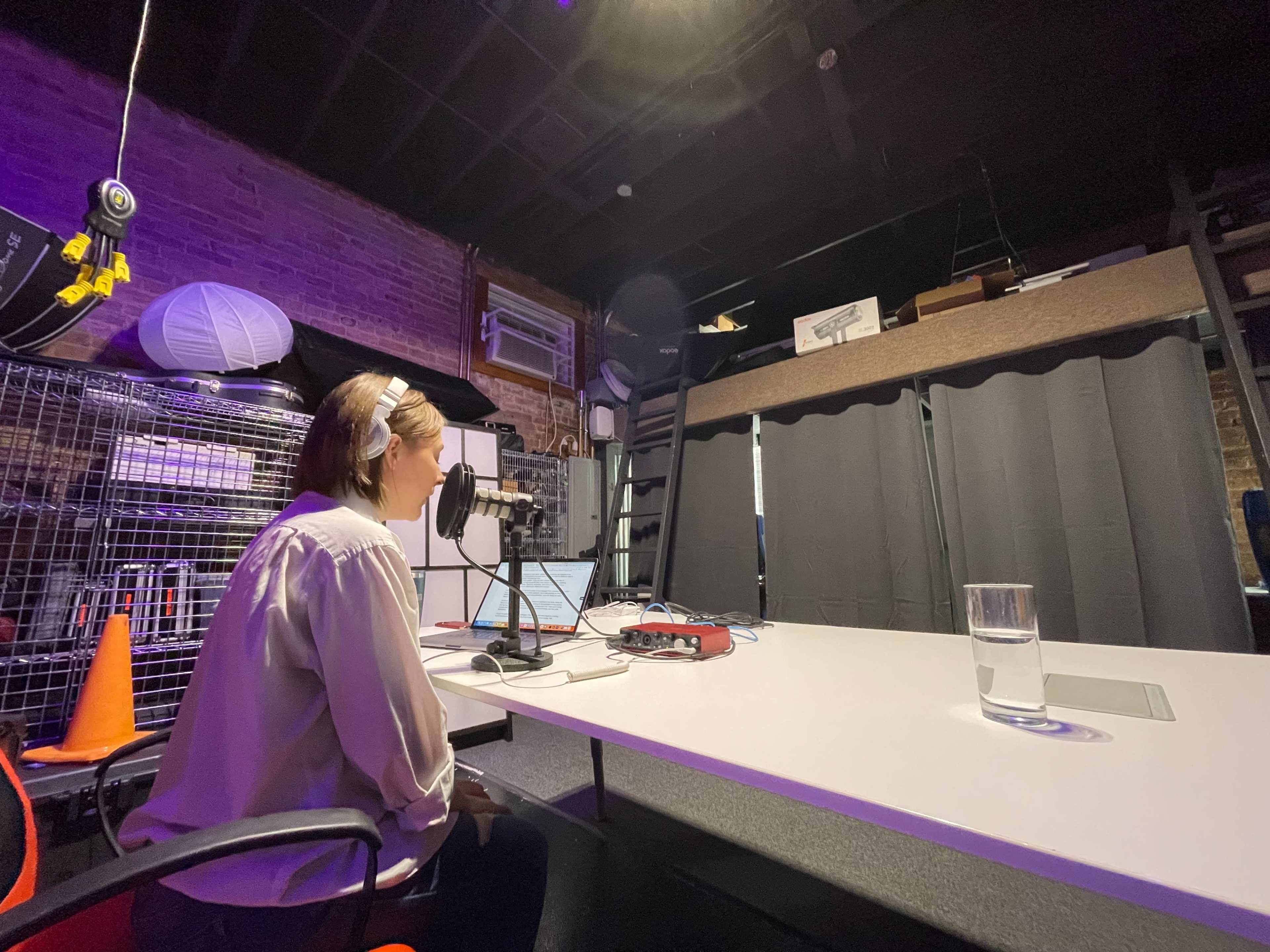 A woman is sitting at a desk with a microphone and laptop, recording in a studio featuring exposed brick walls and a black curtain.