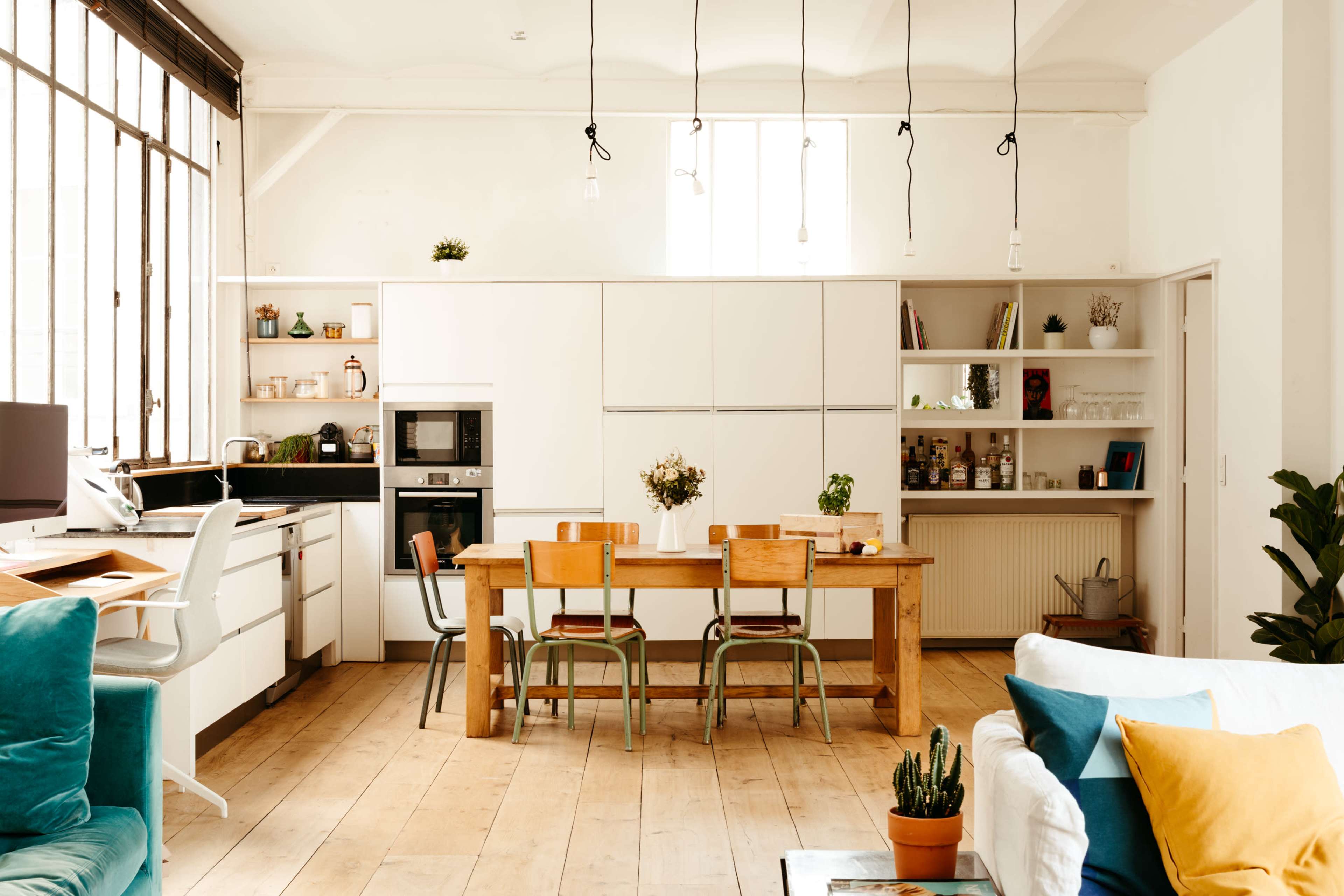 A modern kitchen and dining area features a wooden table surrounded by chairs, with white cabinetry, appliances, and large windows allowing natural light to fill the space.