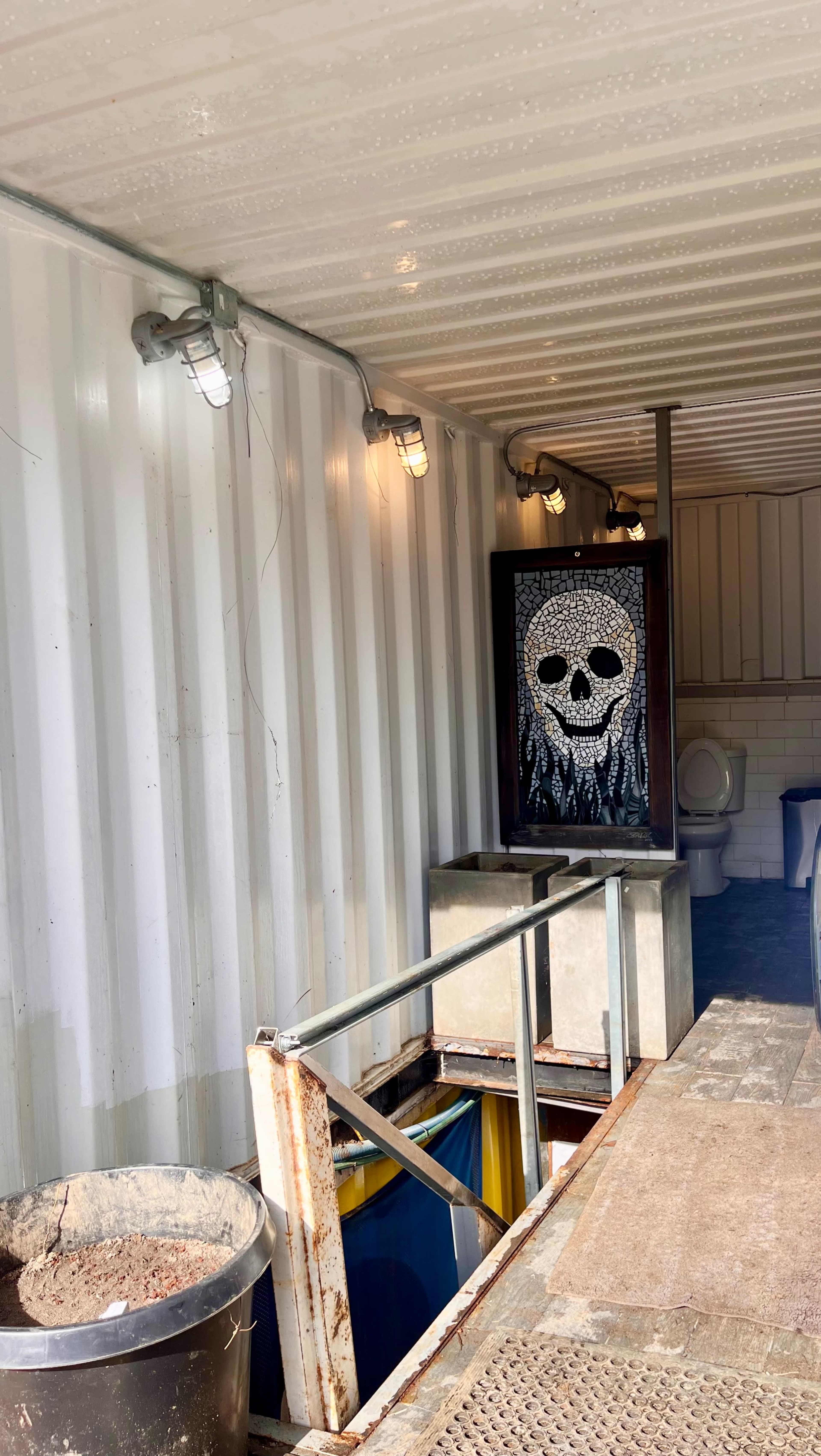 A shipping container interior with a skull mural on a wall, a toilet visible in the background, and two containers on the floor.