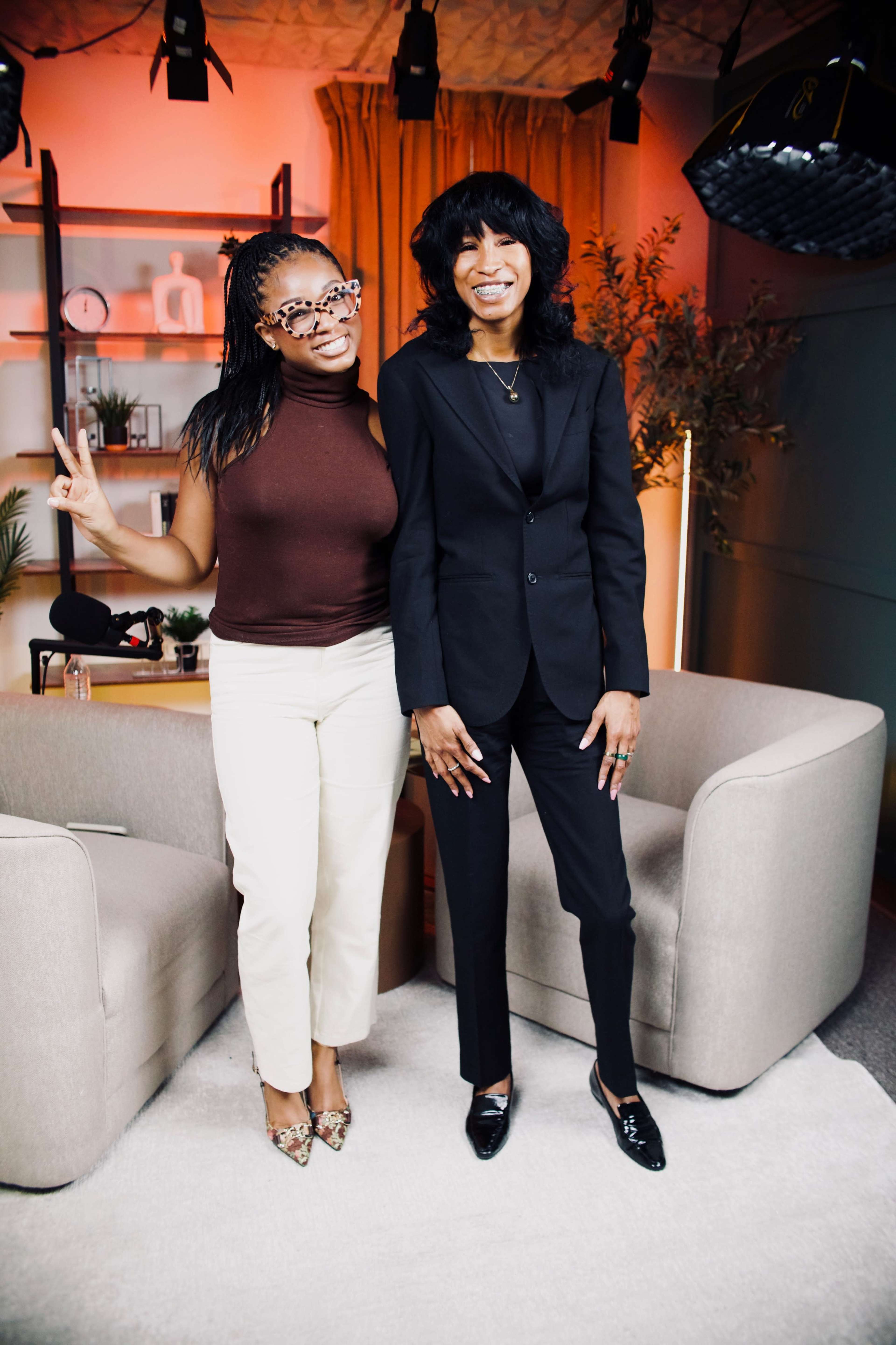 Two women stand together in a well-lit studio setting, one wearing a brown top and cream pants, and the other in a black suit.