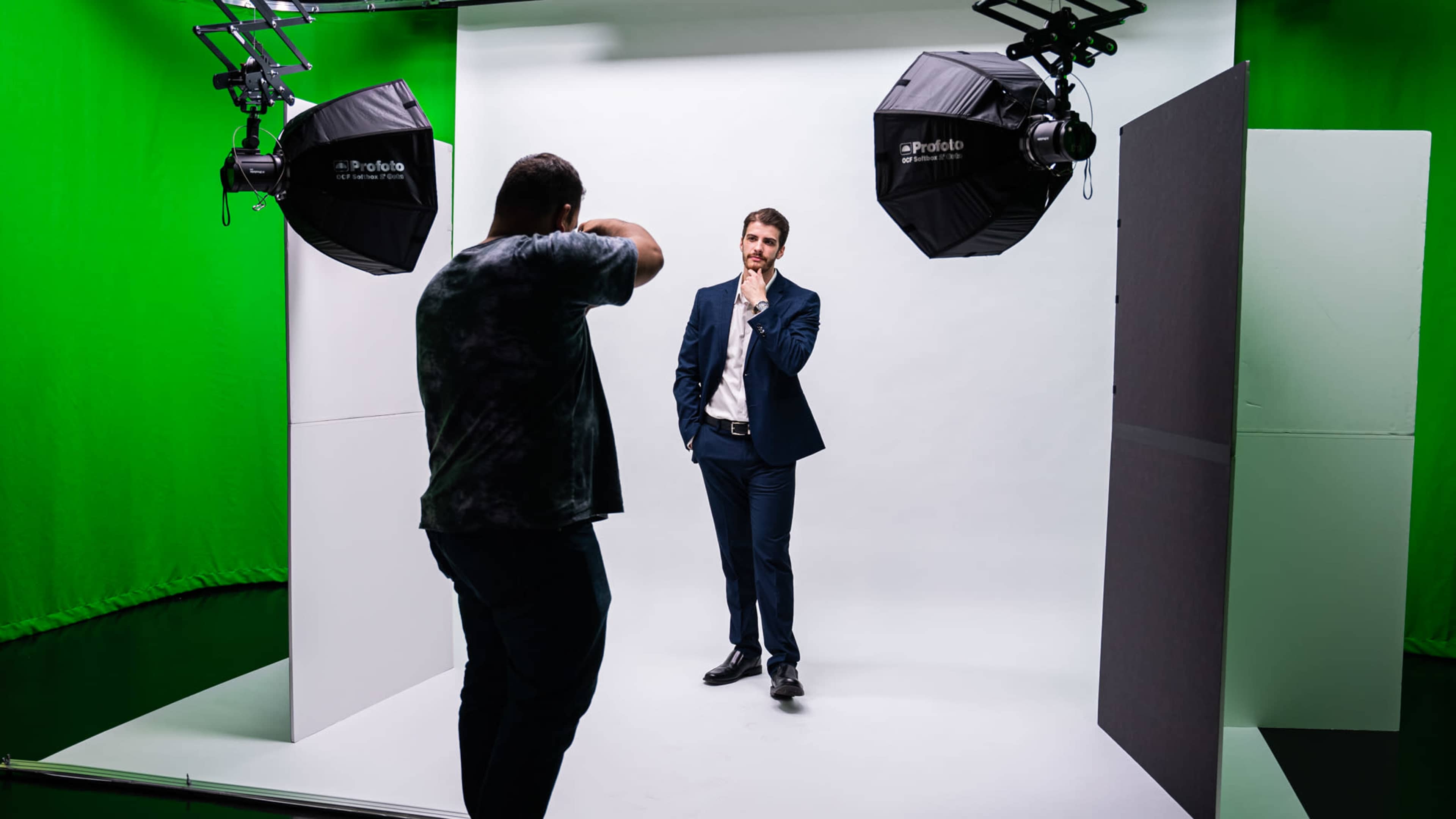 A photographer is taking a picture of a man in a suit against a plain white backdrop illuminated by studio lights in a green screen environment.