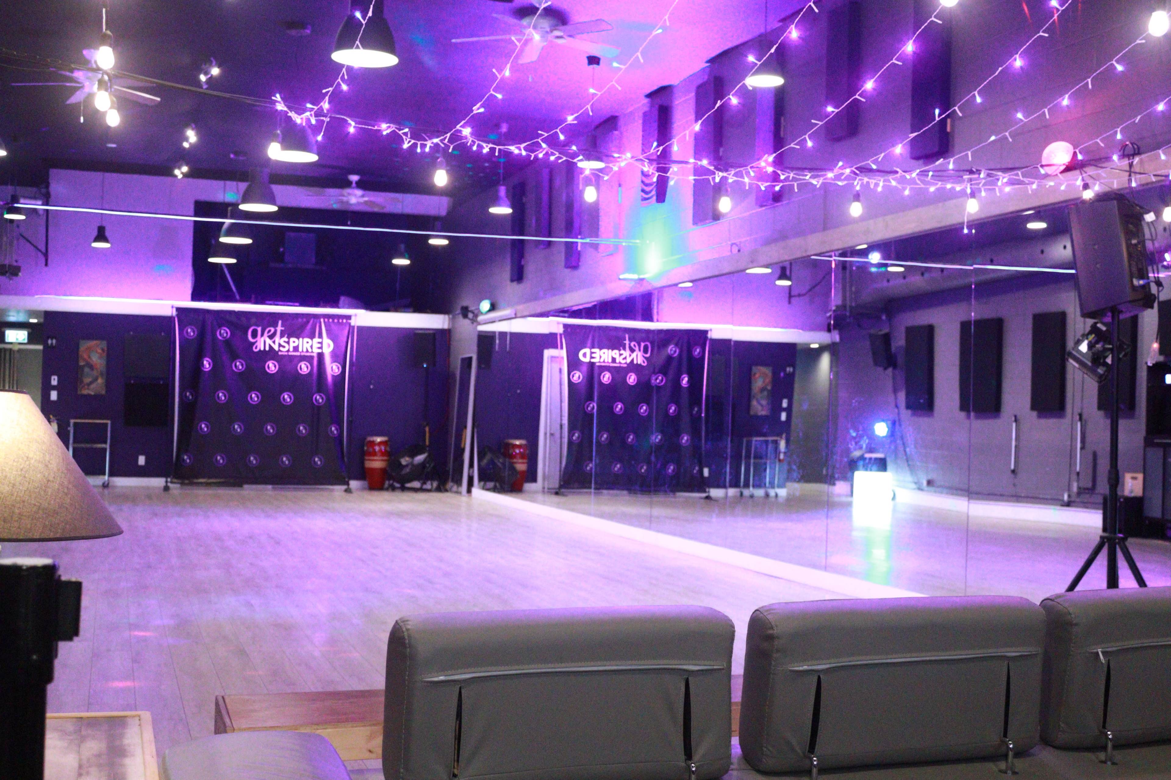 A dance studio with wooden flooring, mirrored walls, and purple lighting, featuring decorative string lights overhead.