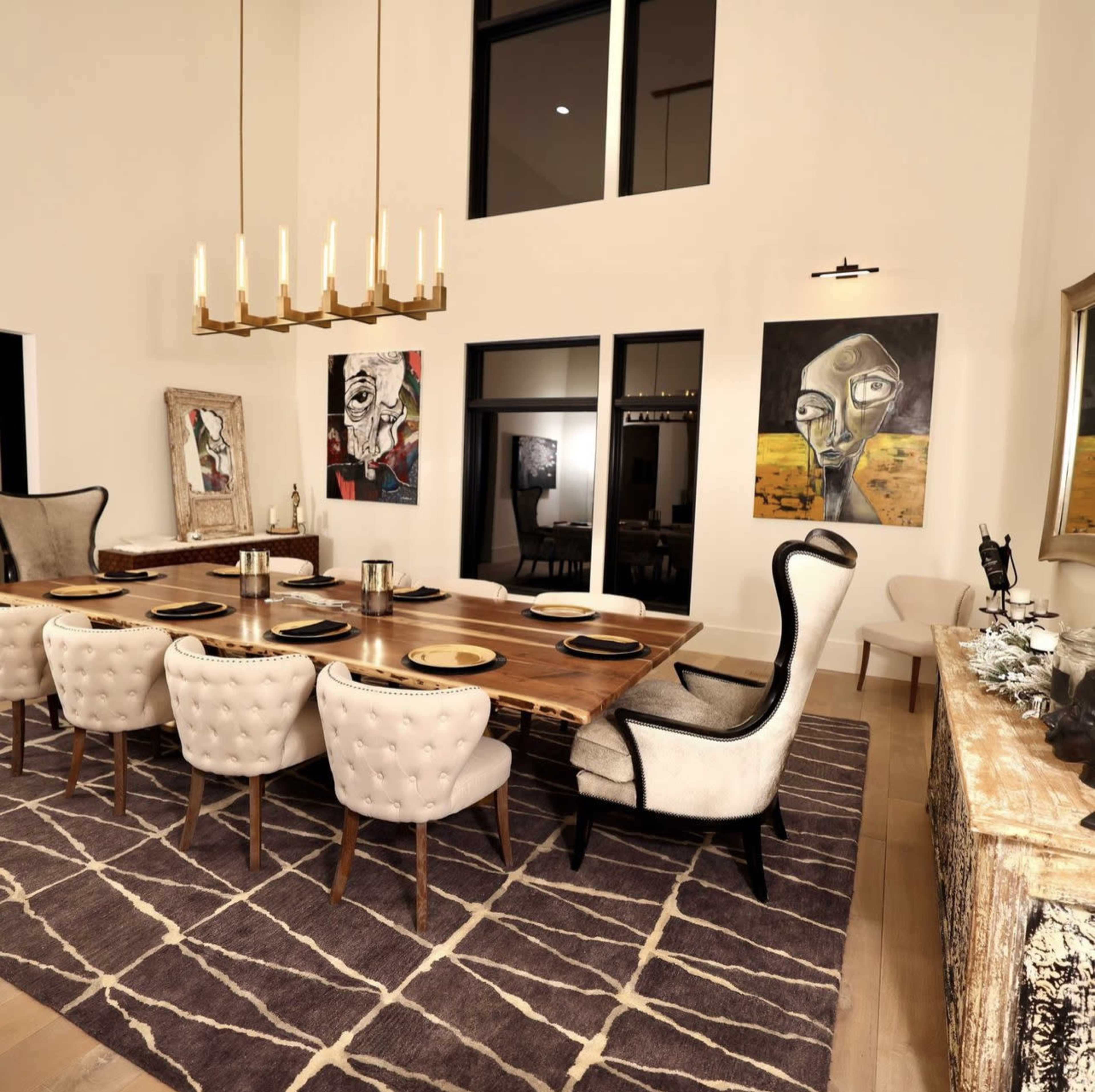 A modern dining room features a long wooden table with upholstered chairs, surrounded by artwork on the walls and illuminated by a chandelier.