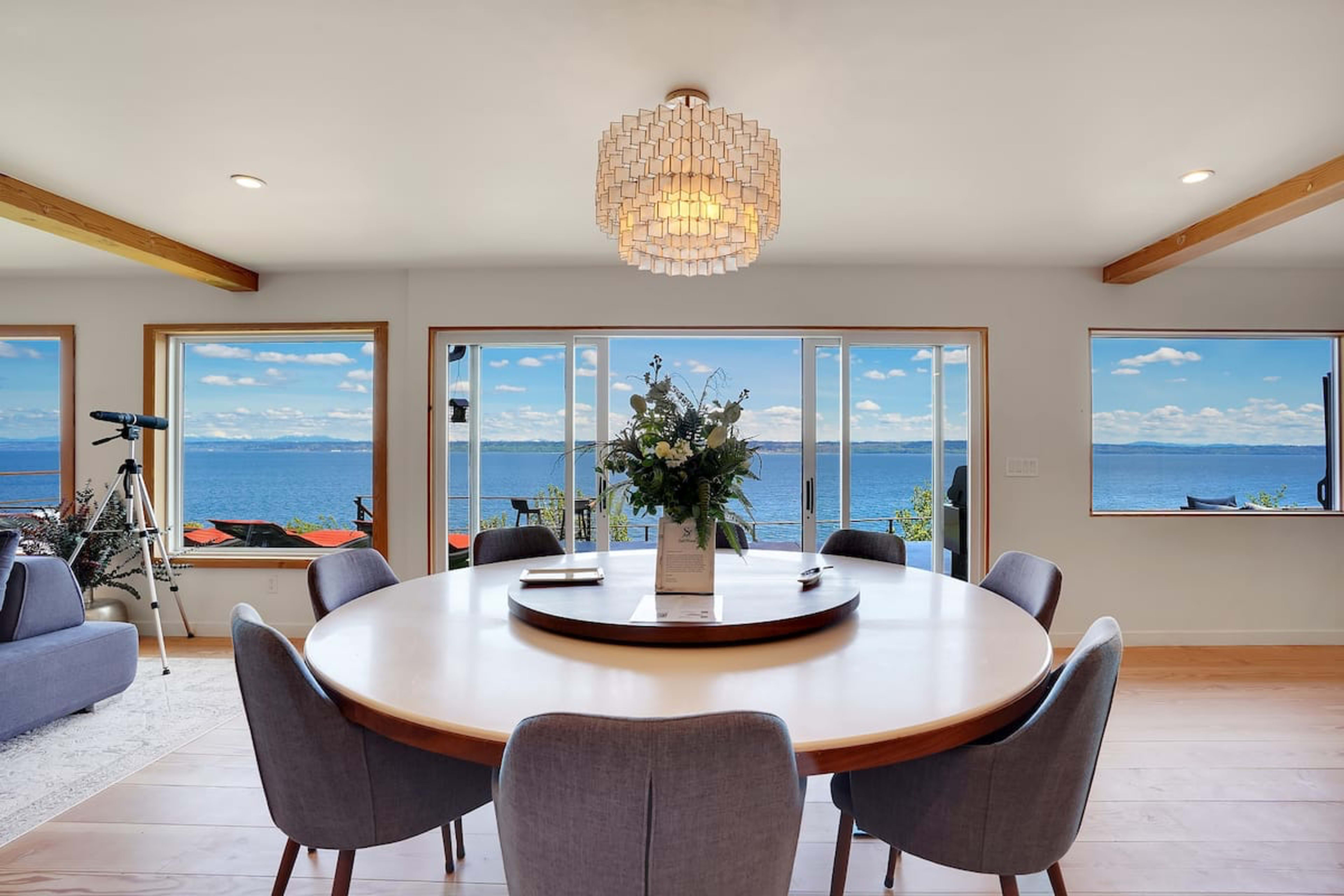A modern dining room features a round table surrounded by gray chairs, with large windows offering a view of the water and sky.