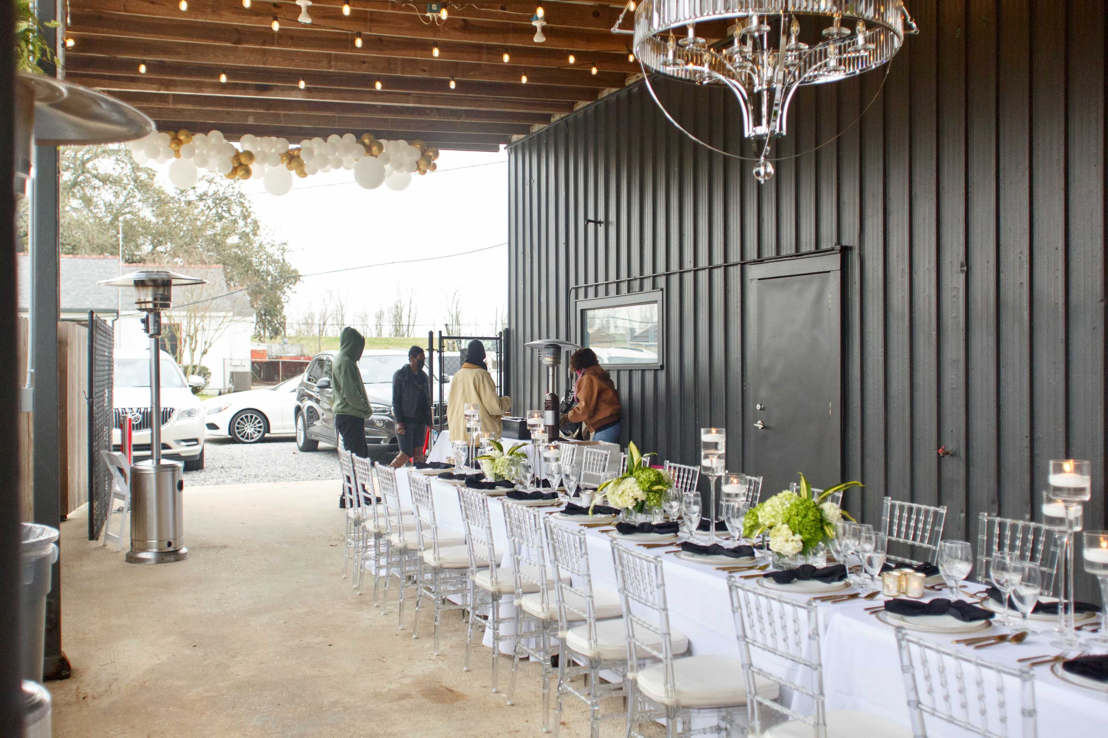 A long, elegantly set dining table with white chairs is arranged under a wooden structure, with people preparing for an event nearby.