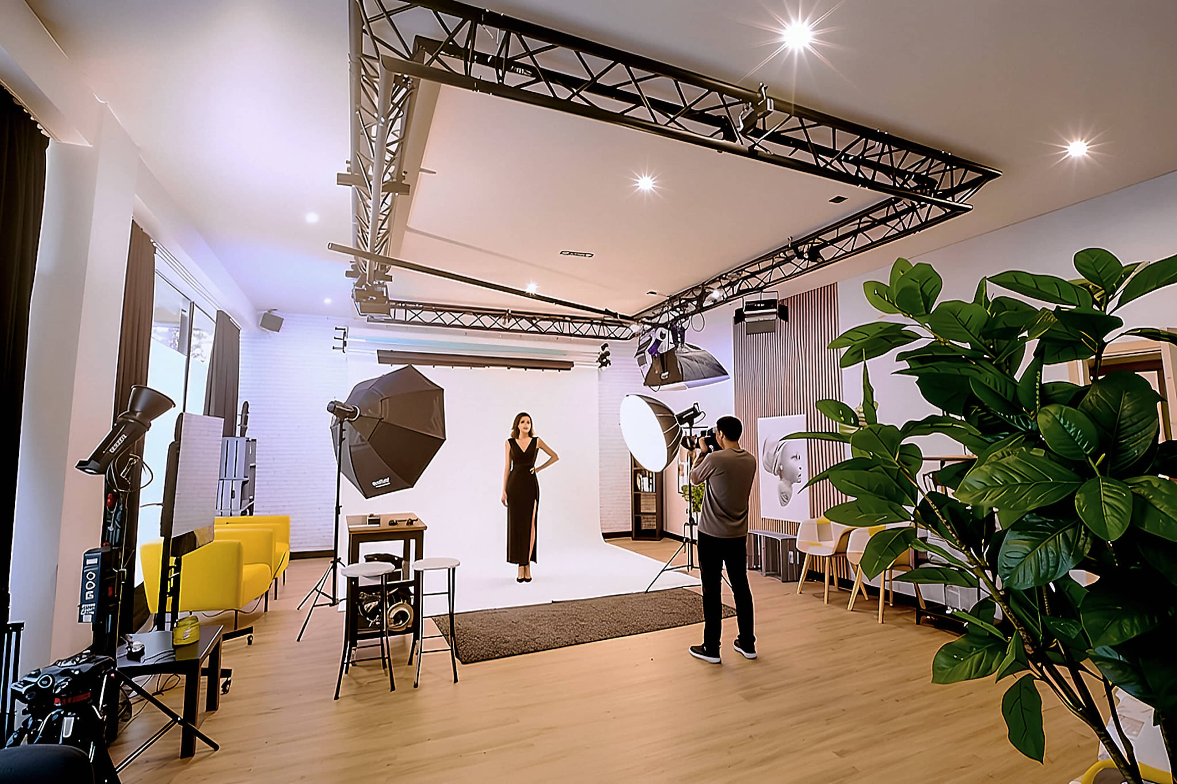 A model poses in a studio while a photographer captures the shot, surrounded by lighting equipment and furniture.