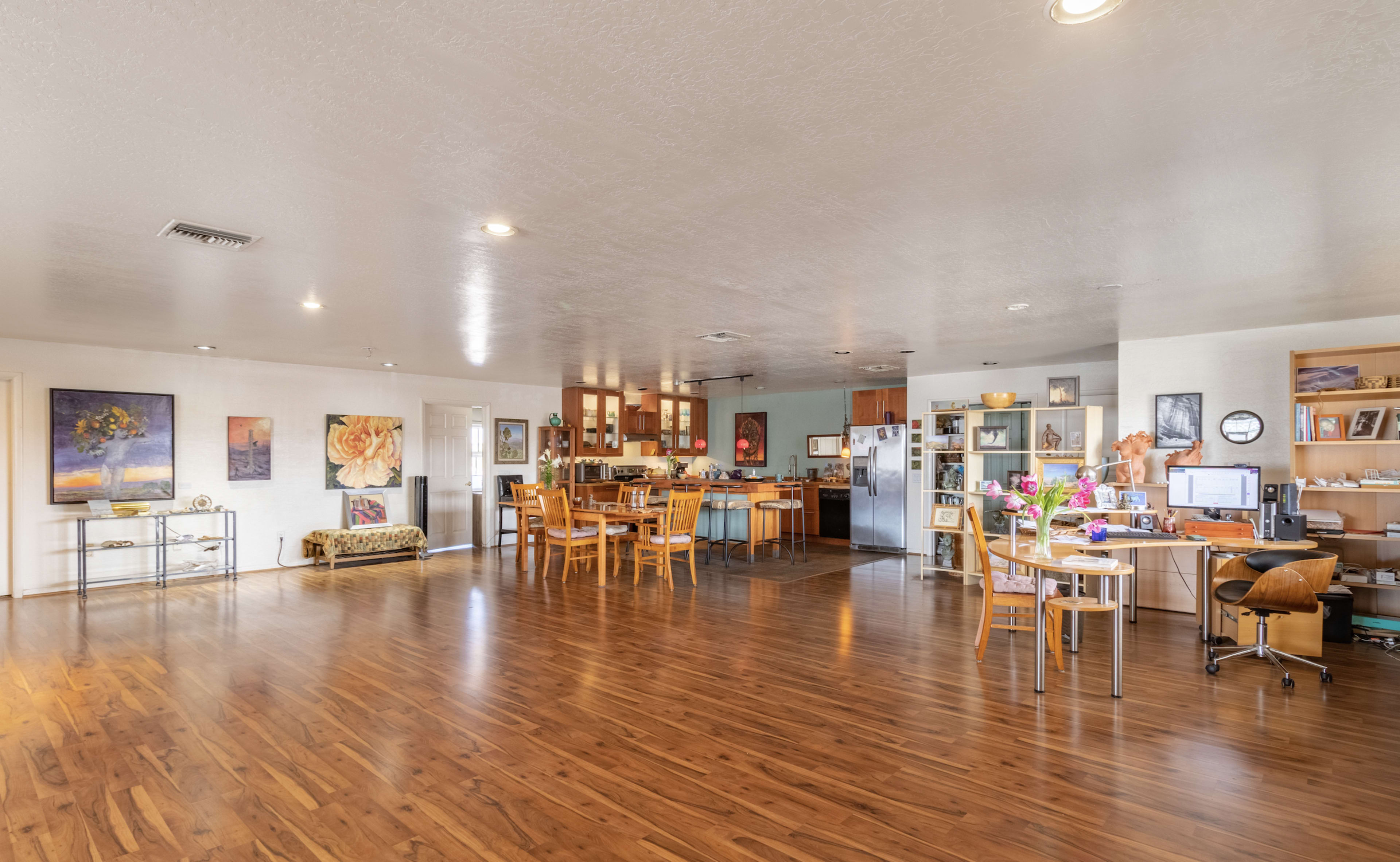 A spacious living area with wooden floors, a dining table, a kitchen, and a workspace surrounded by various artworks on the walls.