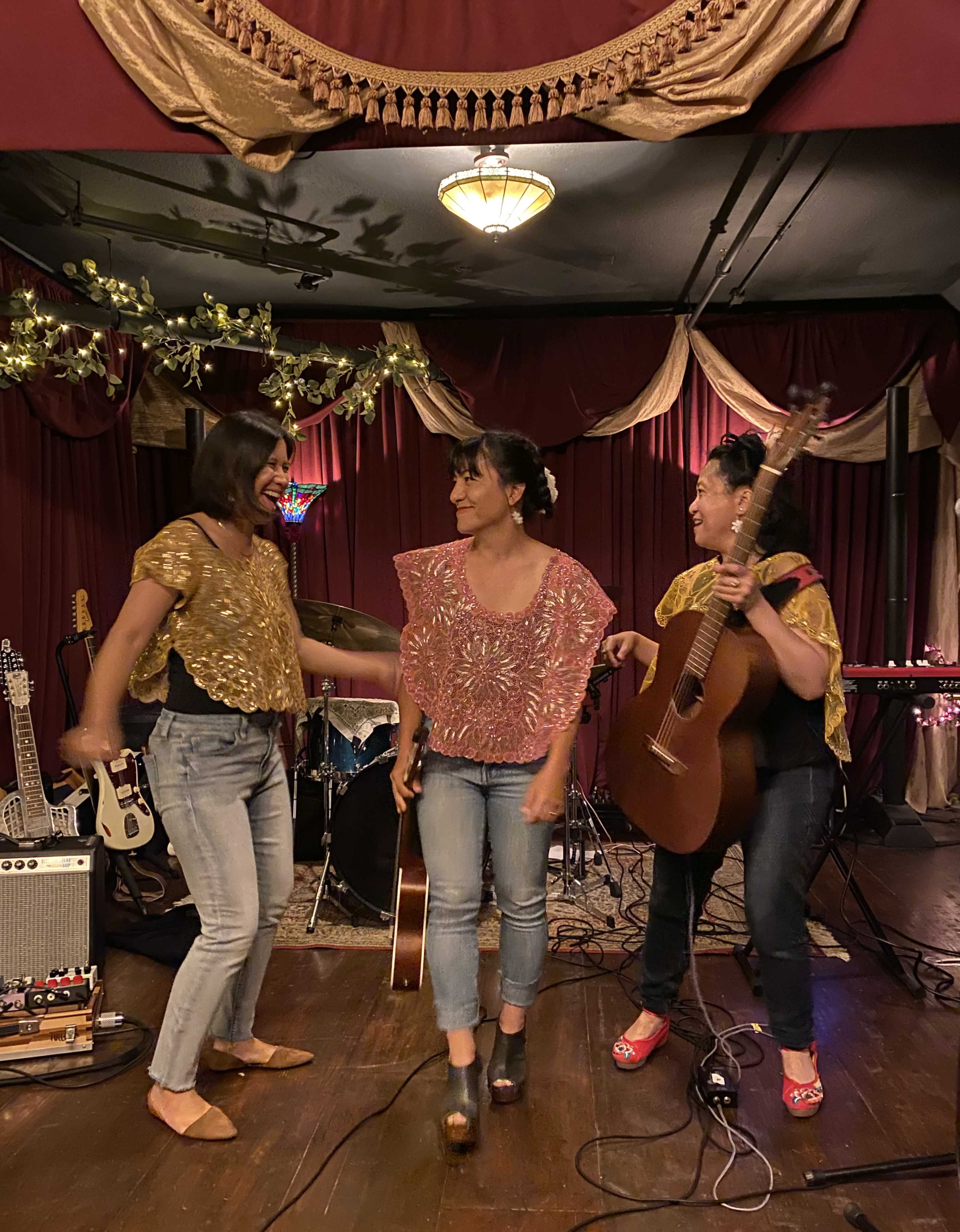 Three women in matching shimmering tops perform on stage, with one playing a guitar and the other two dancing in front of a band setup.