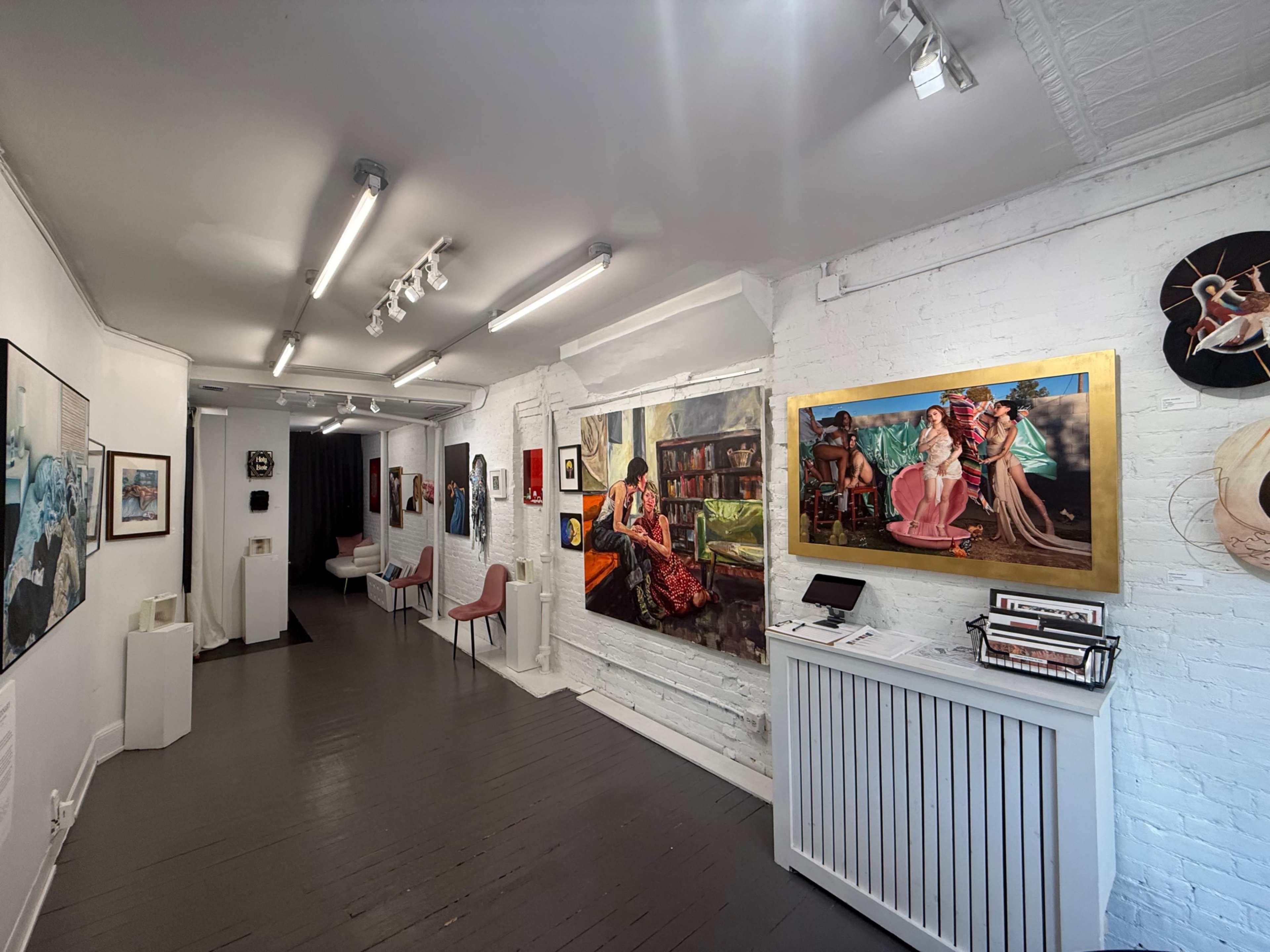 The image shows an art gallery with white brick walls displaying several paintings, while a corridor leads to additional artworks and seating areas.