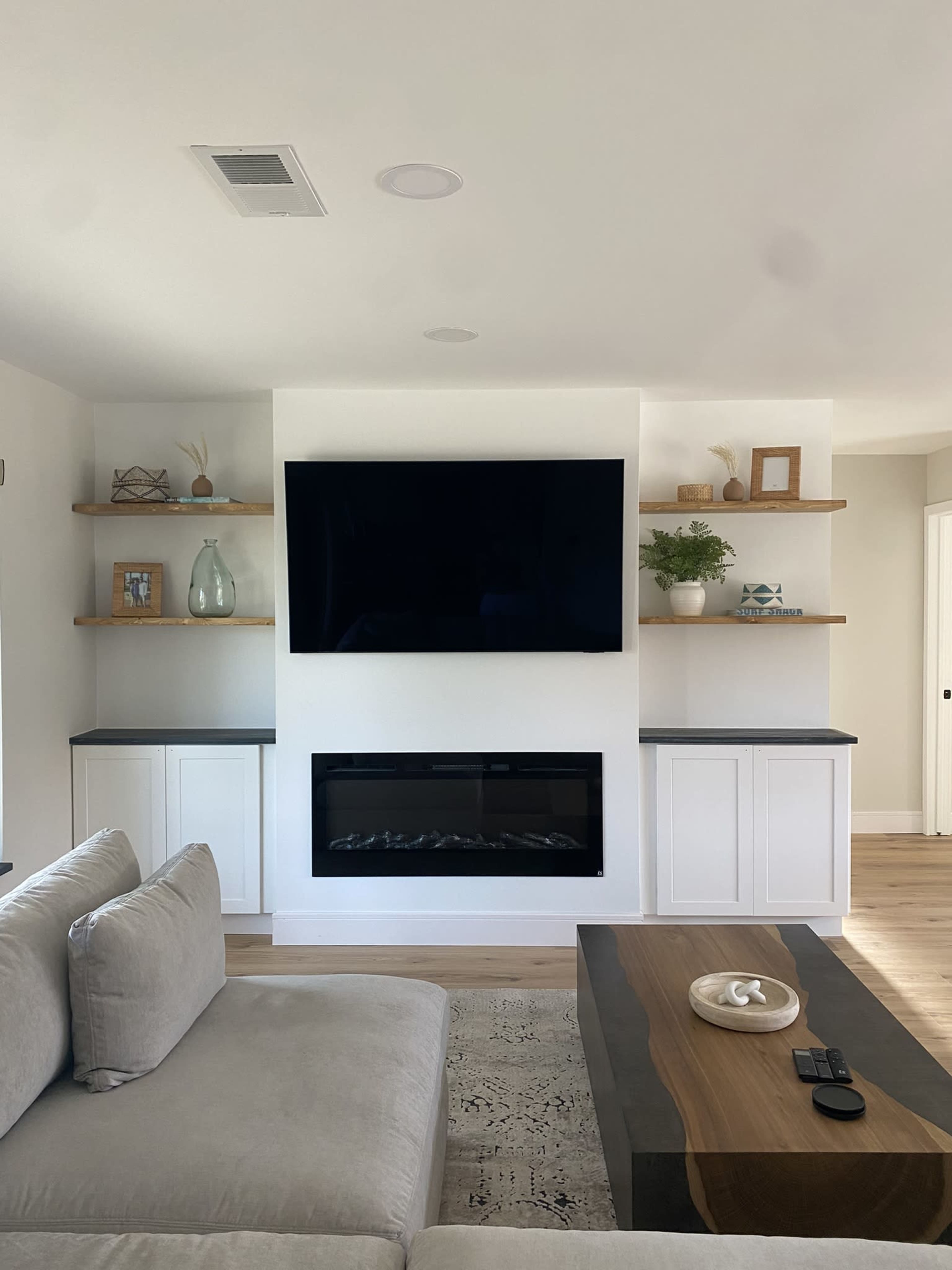 The living room features a modern setup with a flat-screen TV mounted above a fireplace, flanked by open shelves and a beige sofa.