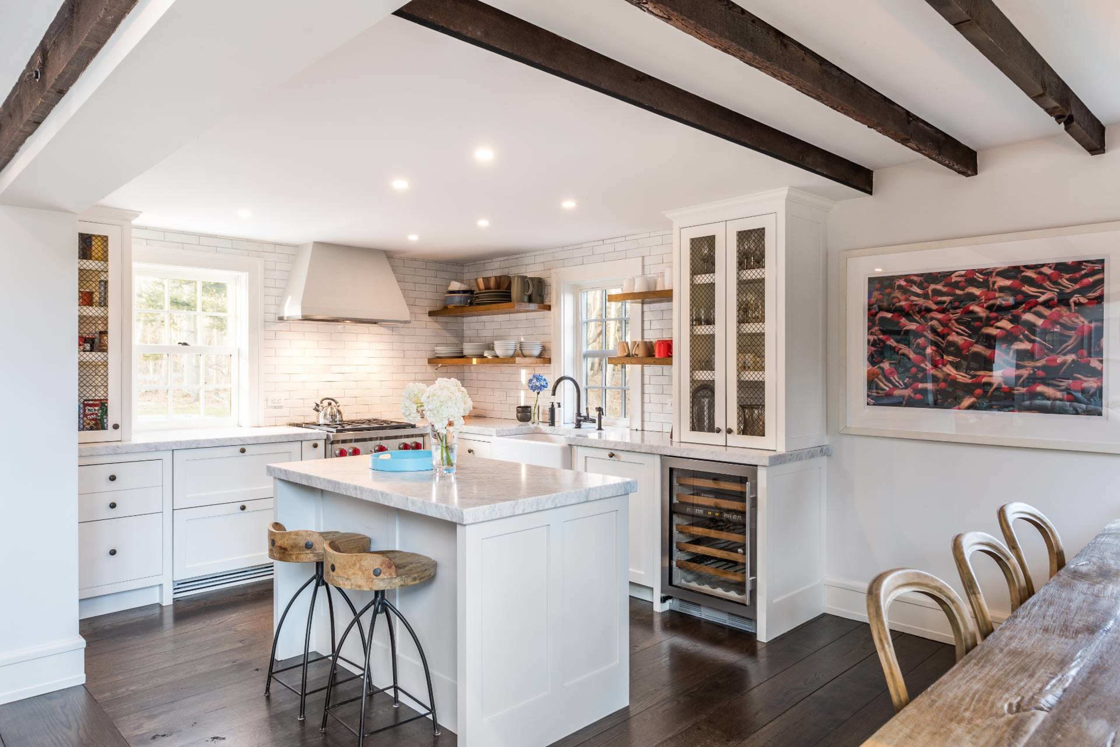 A modern kitchen features white cabinetry, a large island with seating, and exposed wooden beams overhead.
