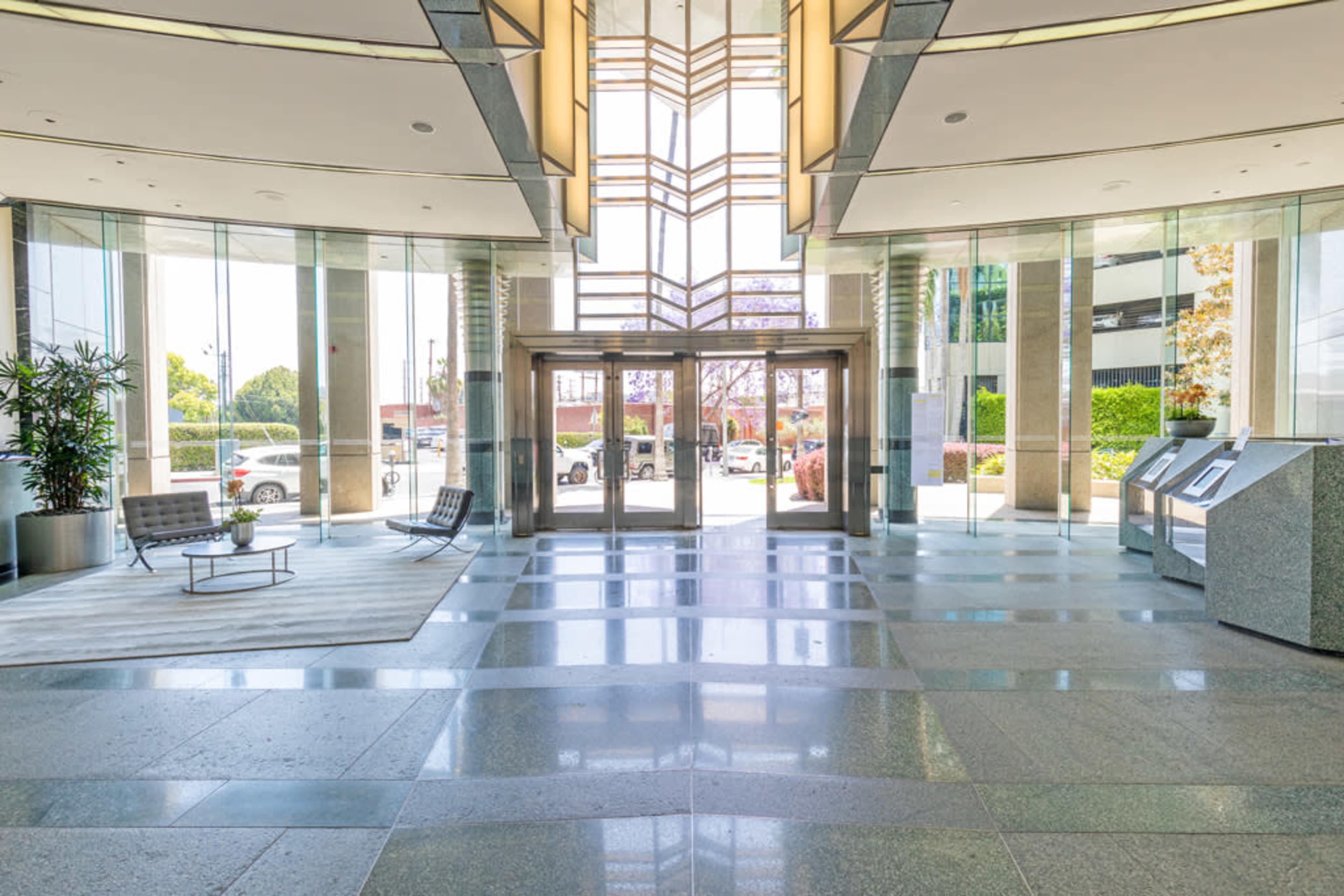 A modern lobby with large glass doors, geometric lighting, and polished stone floors, alongside seating areas and informational kiosks.