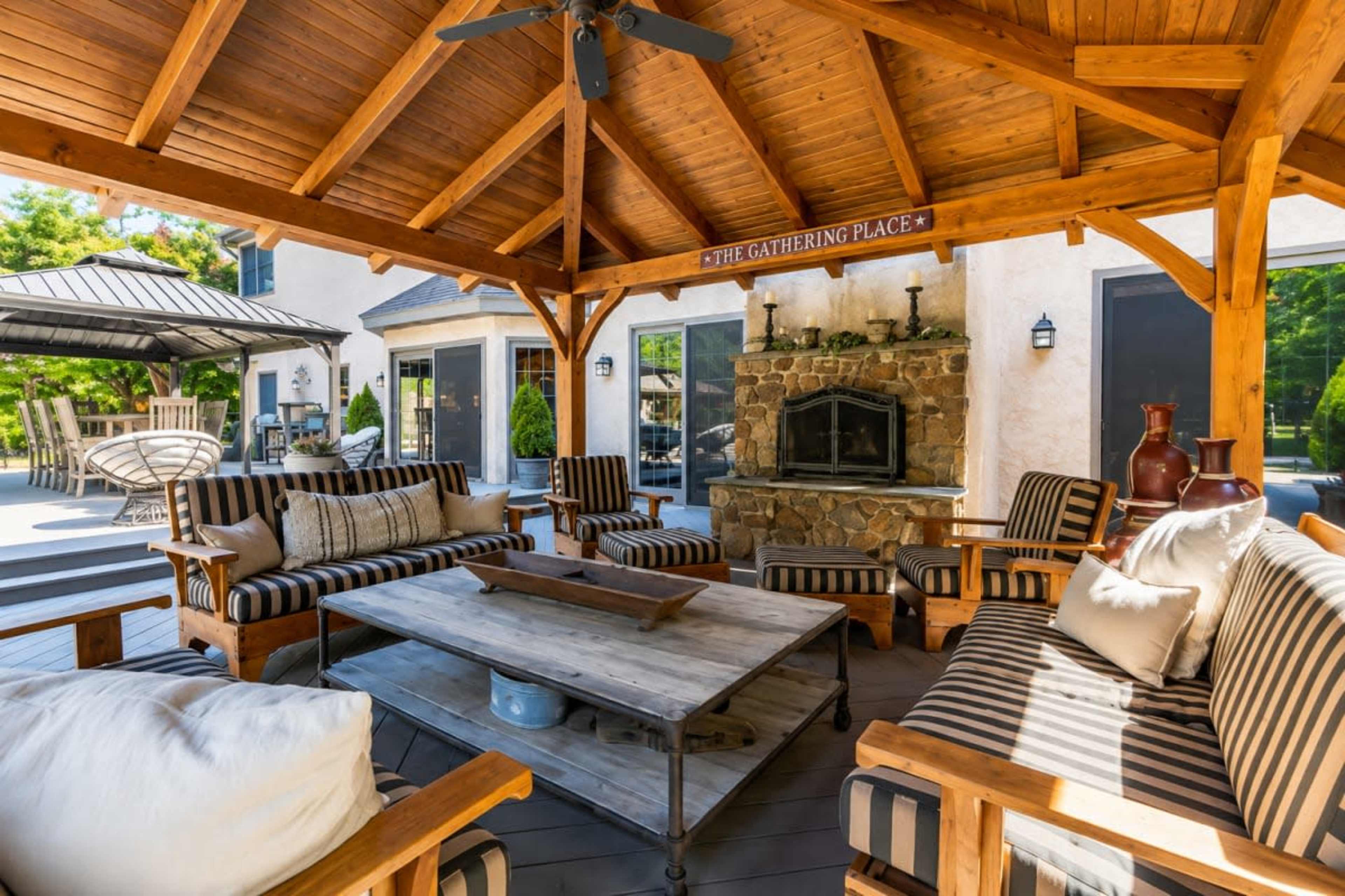 The image shows a wooden gazebo with a stone fireplace, surrounded by seating arrangements including striped chairs and a central wooden table.