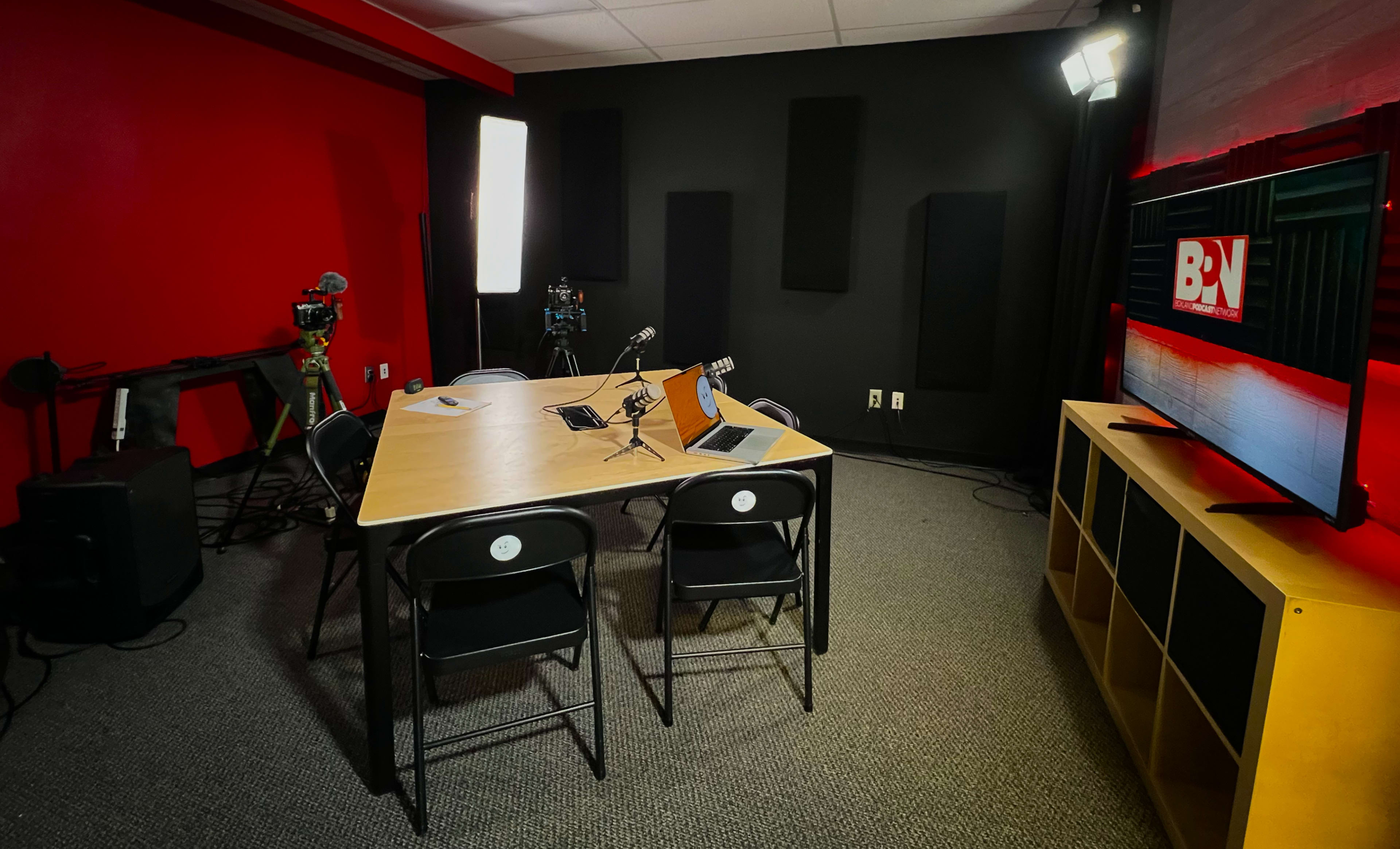 A modern recording studio with a central table, two chairs, cameras on tripods, and soundproofing panels on the walls.