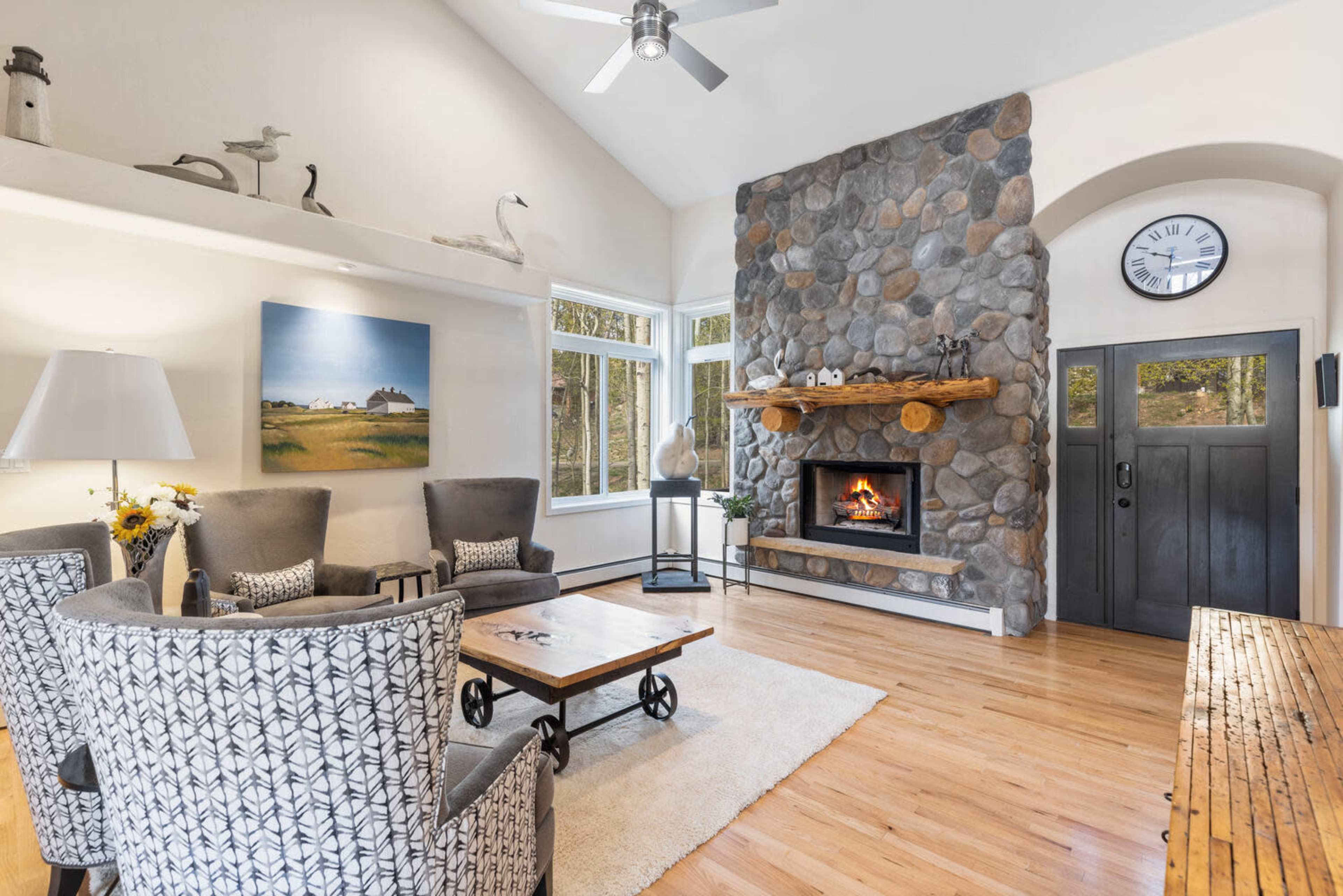The living room features a stone fireplace, two armchairs, a wooden coffee table, and large windows allowing natural light to illuminate the space.