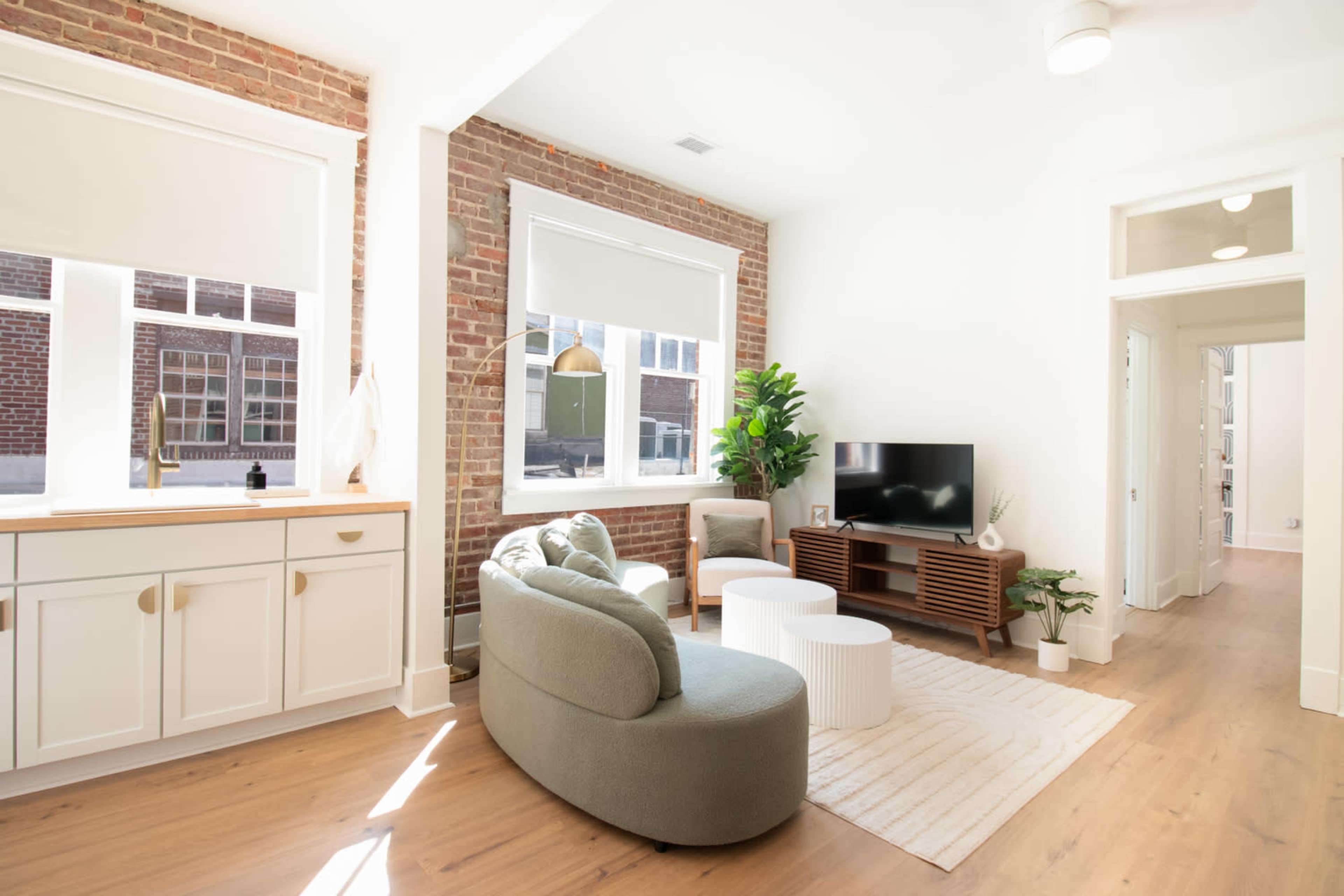 A modern living room featuring a curved green sofa, round coffee tables, a television on a wooden stand, and large windows with natural light illuminating the space.