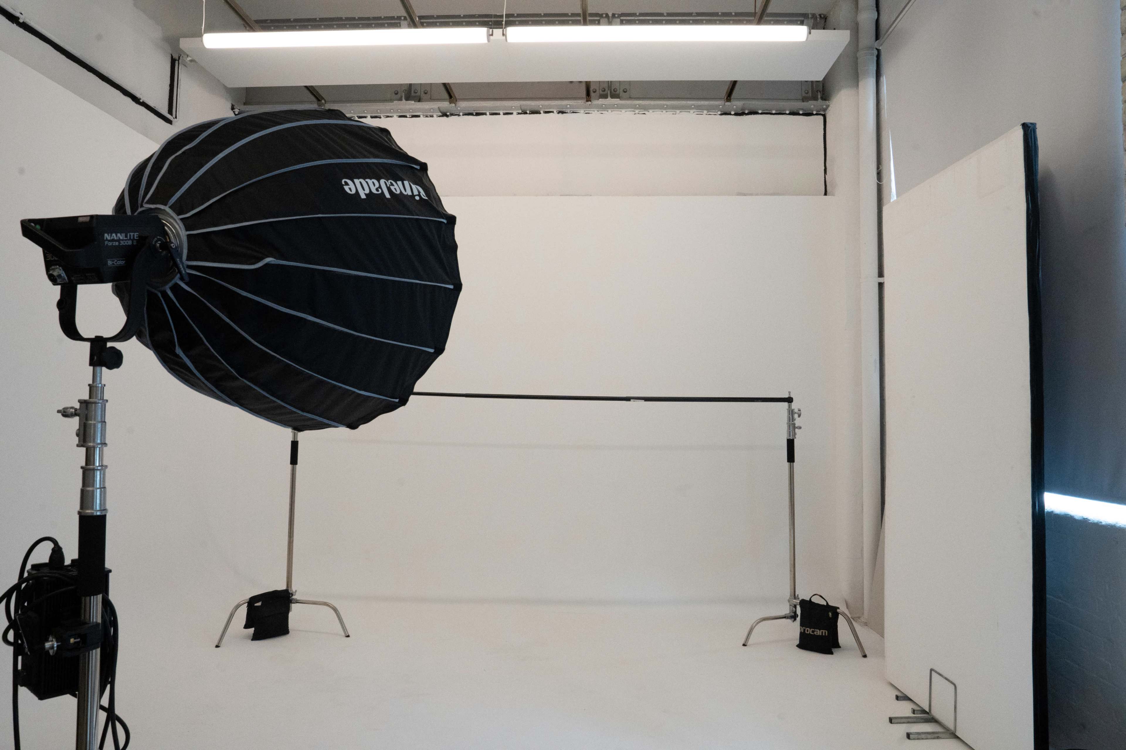 The image shows a photography studio setup with a large softbox light on a stand, a backdrop support system, and blank white walls.