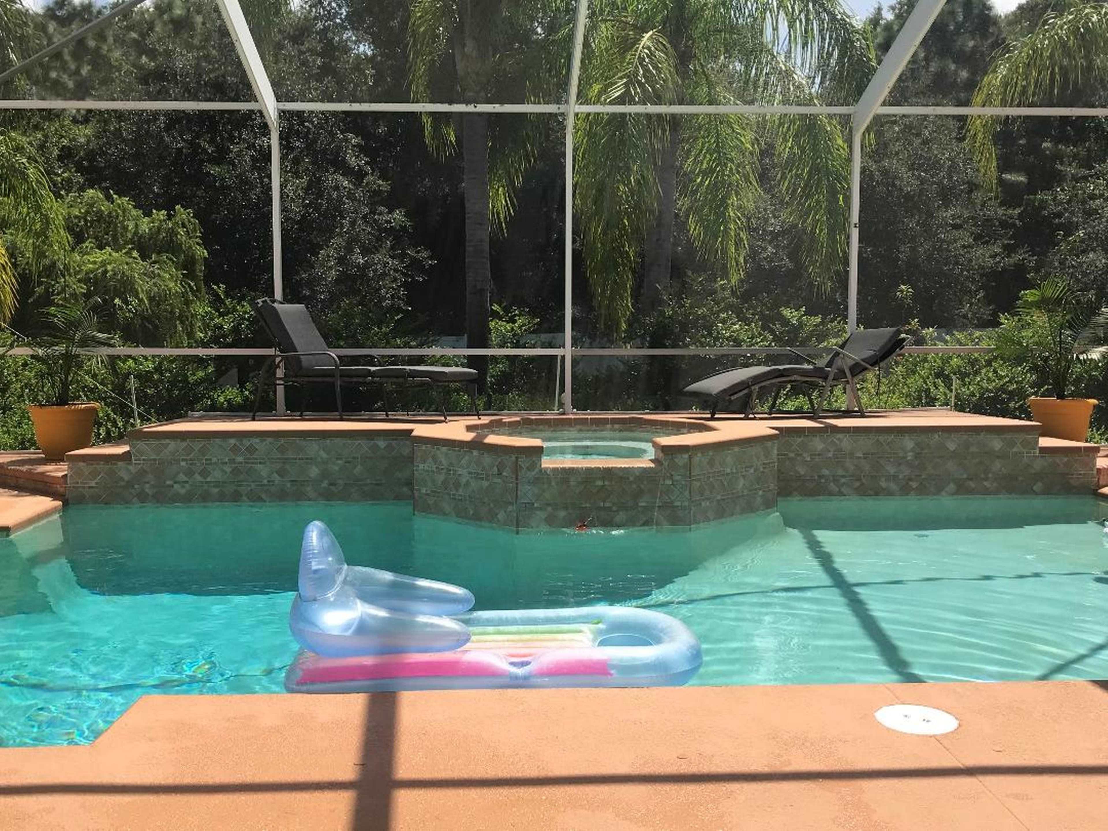 A swimming pool with a floating lounge, surrounded by patio chairs and lush greenery visible through a screened enclosure.