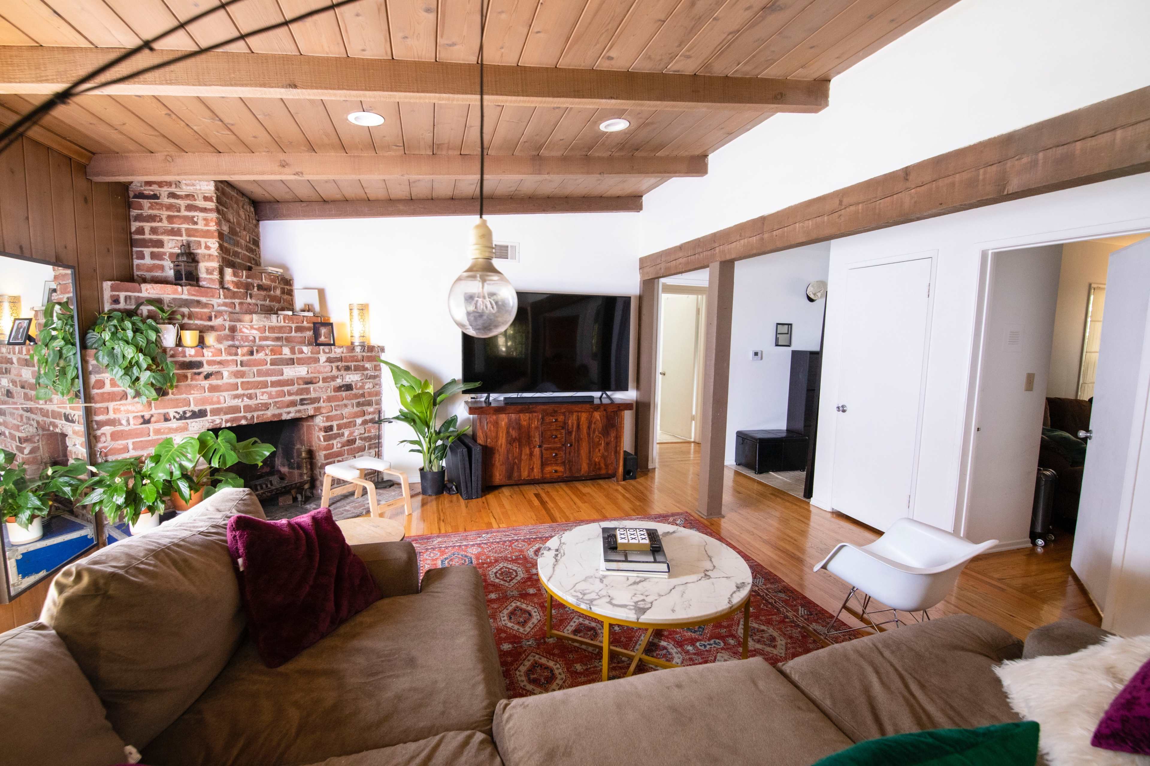 The living room features a brick fireplace, wooden beams, and a television, with a marble coffee table and a patterned rug.