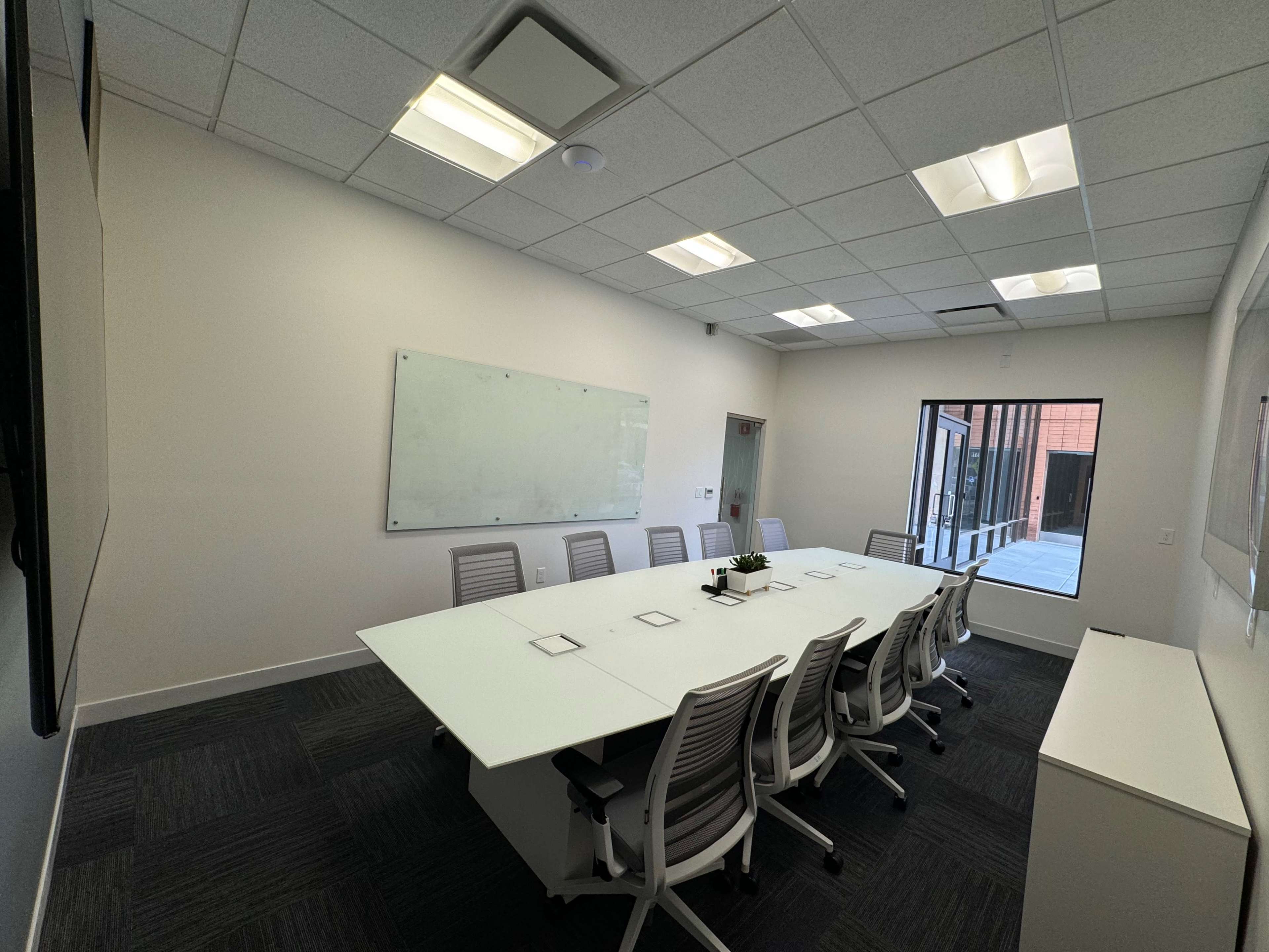 A modern conference room with a long white table, several ergonomic chairs, and a large window providing natural light.