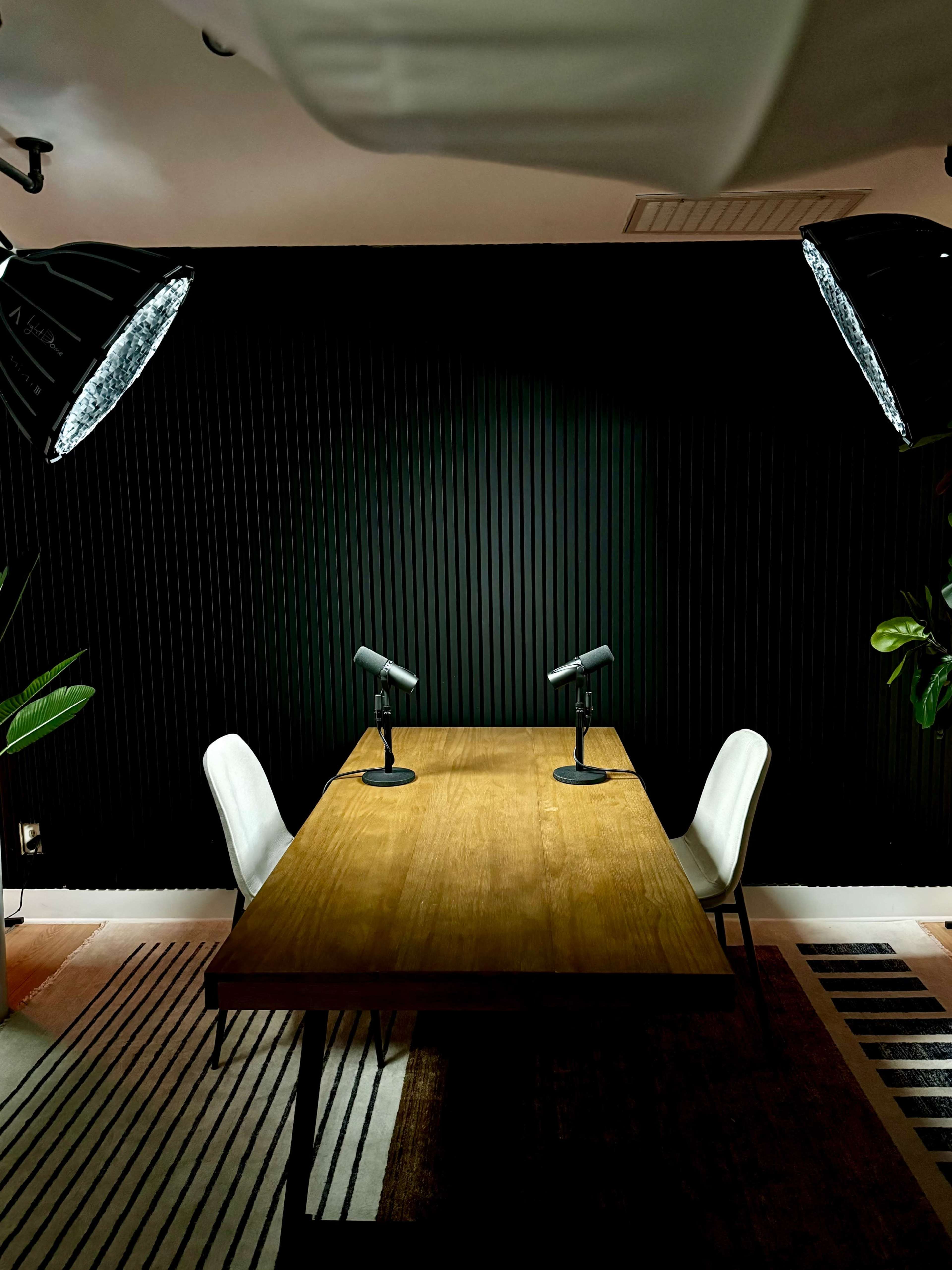 A wooden table with two chairs and microphones is set under bright lights against a dark, ribbed wall.