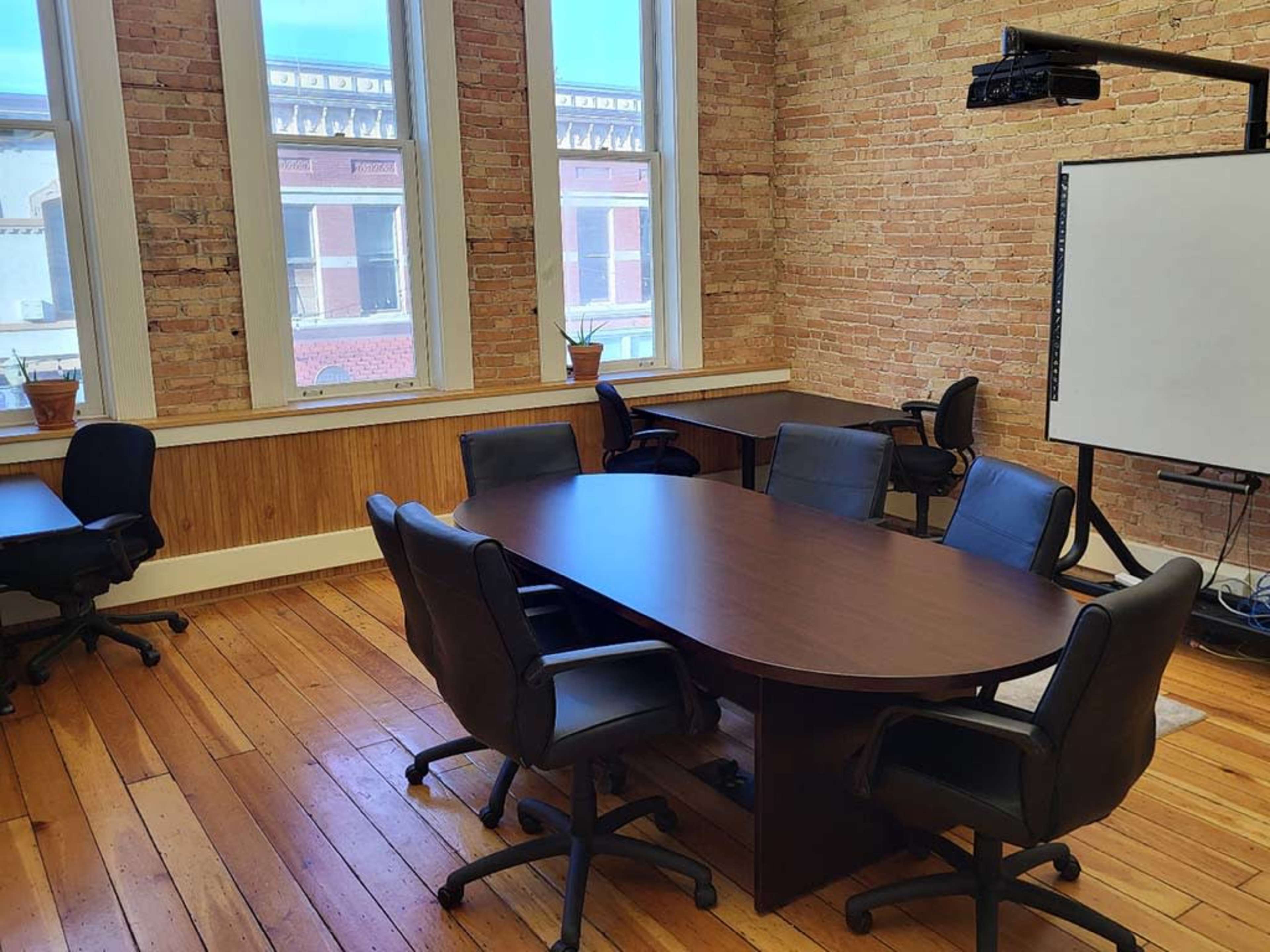 A conference room features a large wooden table surrounded by eight black chairs, with a presentation screen and windows letting in natural light.