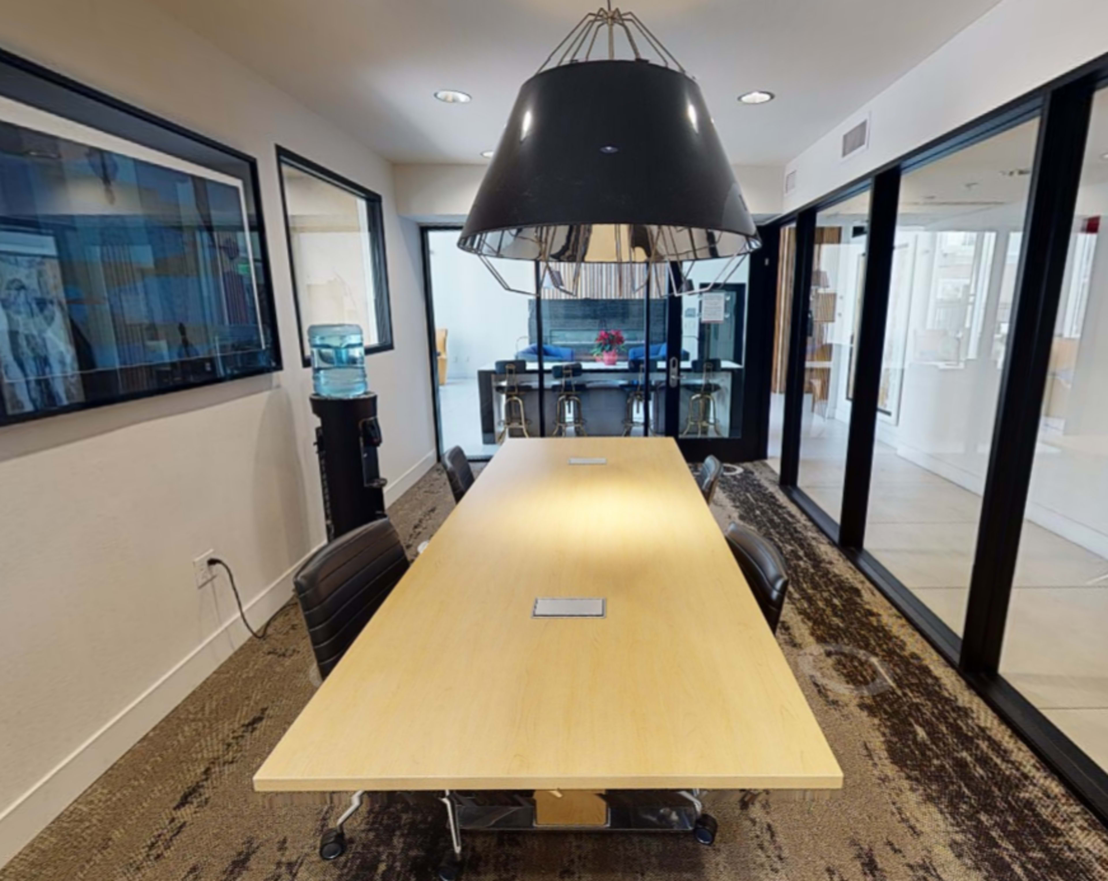 A long wooden conference table is illuminated by a large pendant light in a modern meeting room with glass walls and a water cooler.