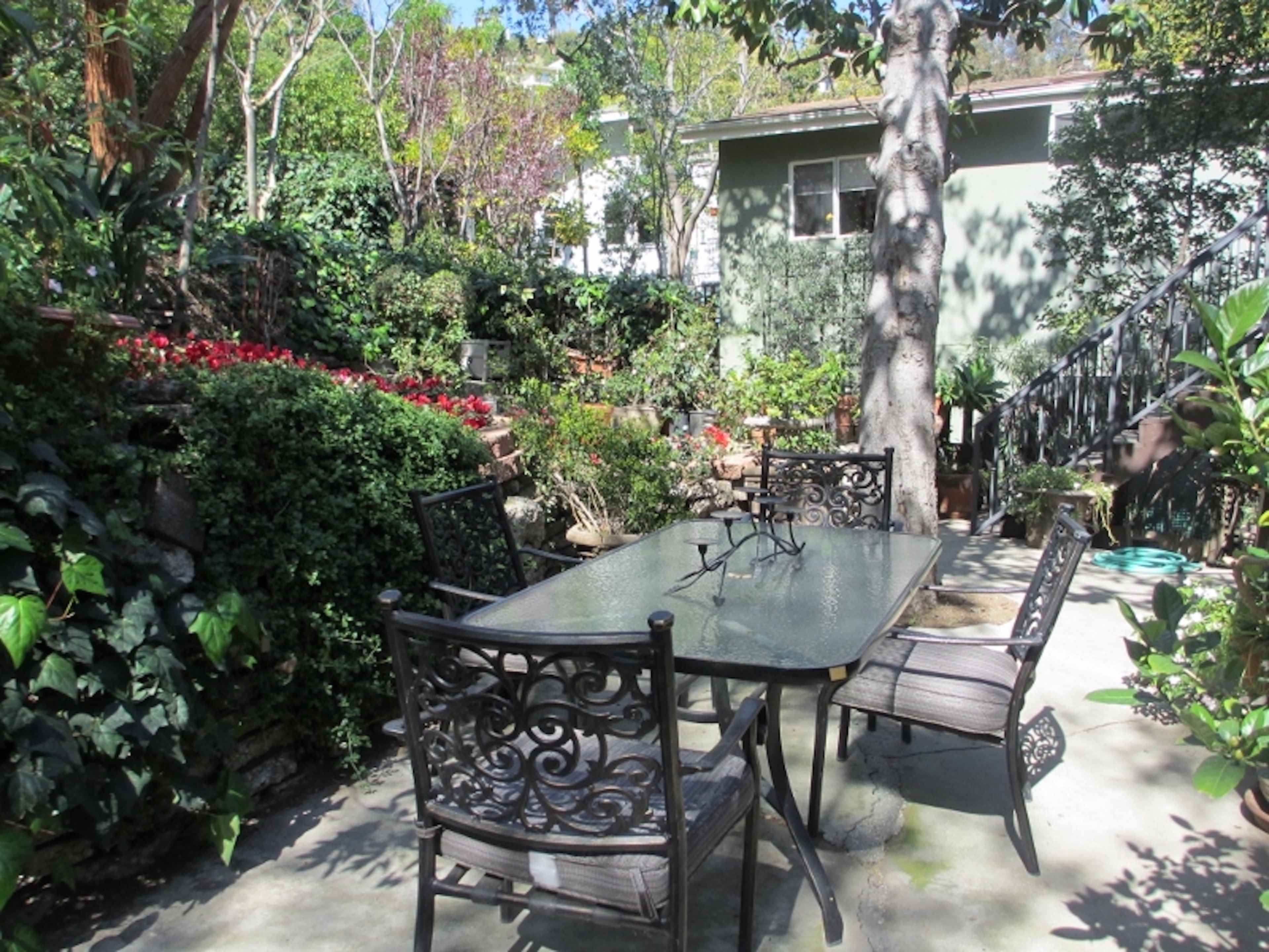 A set of four wrought iron chairs surrounds a glass-top table in a lush garden filled with greenery and flowers.
