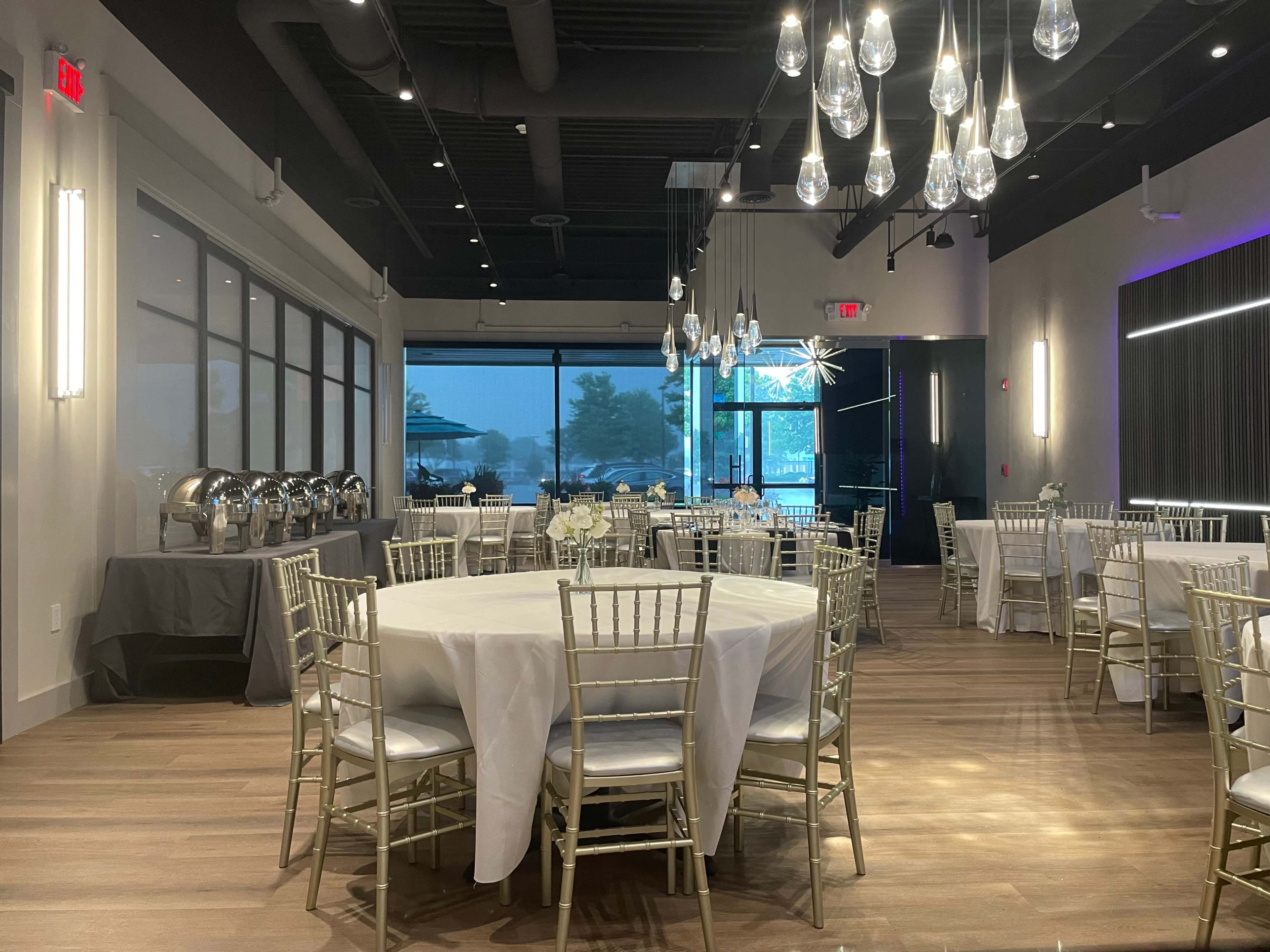 An elegantly set dining area with round tables, silver chairs, and a buffet setup, illuminated by modern pendant lights, and large windows revealing a cloudy exterior.