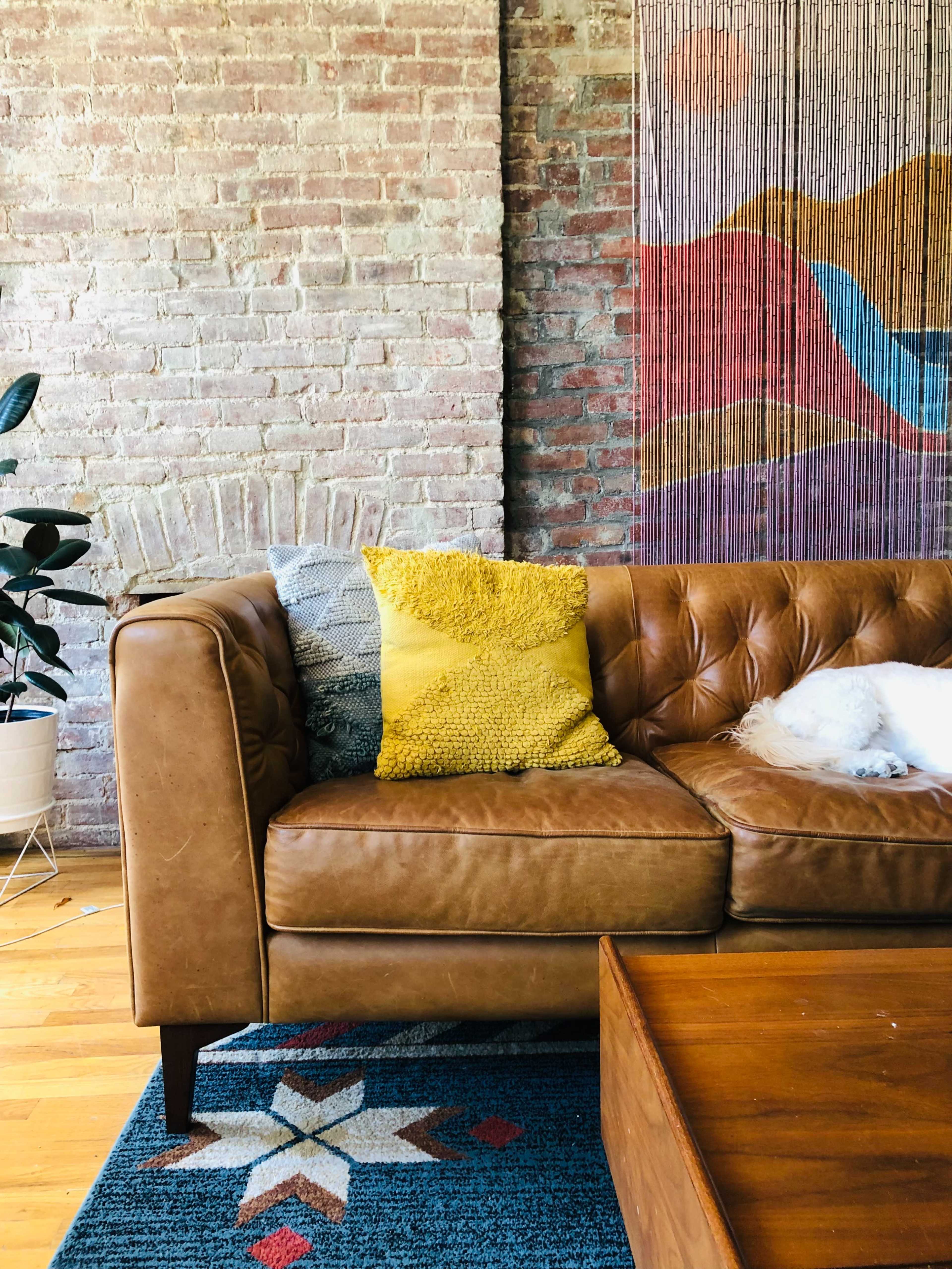 A leather couch with a yellow pillow is positioned in front of a wooden coffee table against a brick wall adorned with colorful artwork.