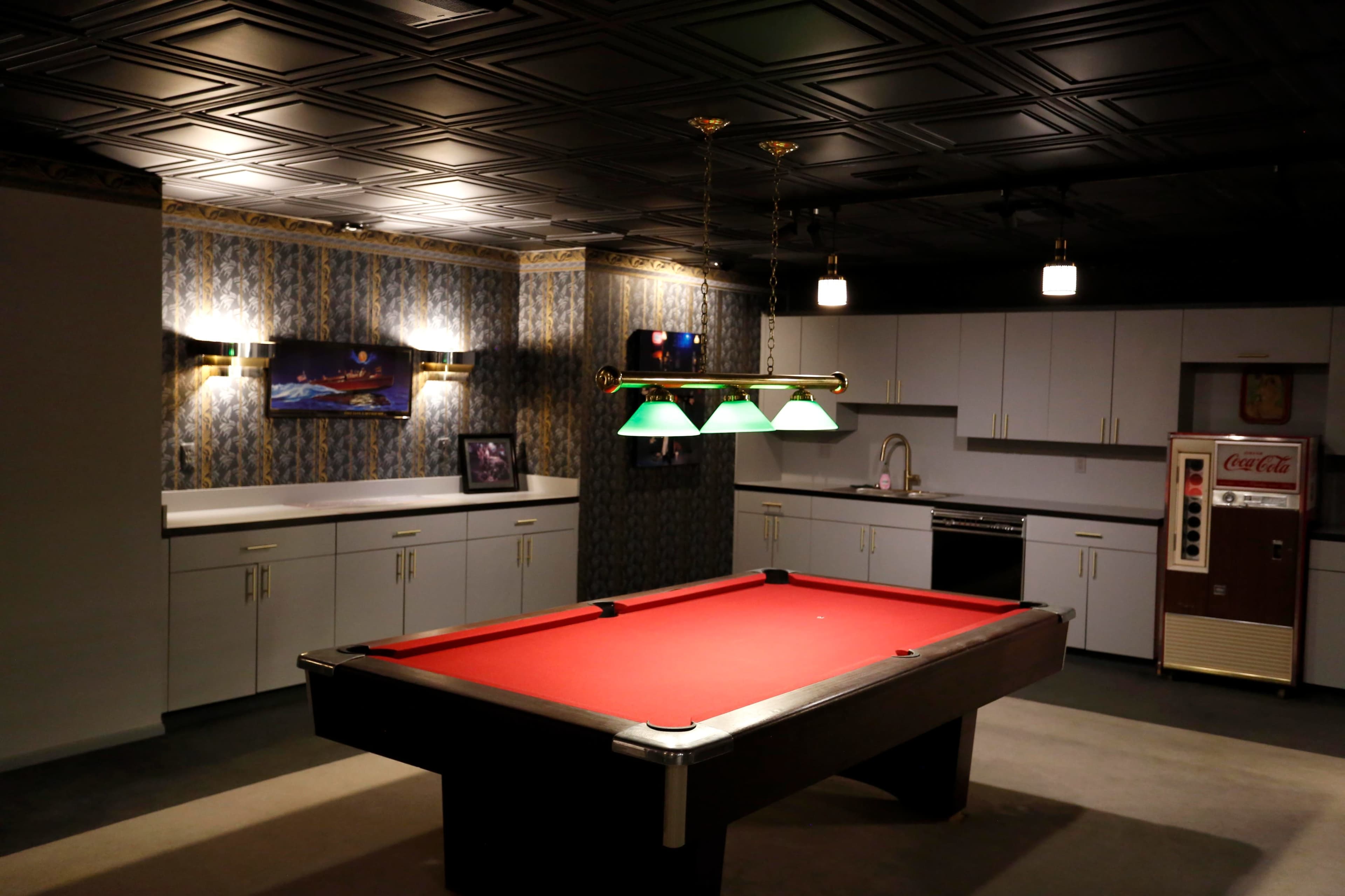 A well-lit game room featuring a red billiards table, a vintage Coca-Cola vending machine, and a kitchenette with white cabinetry.