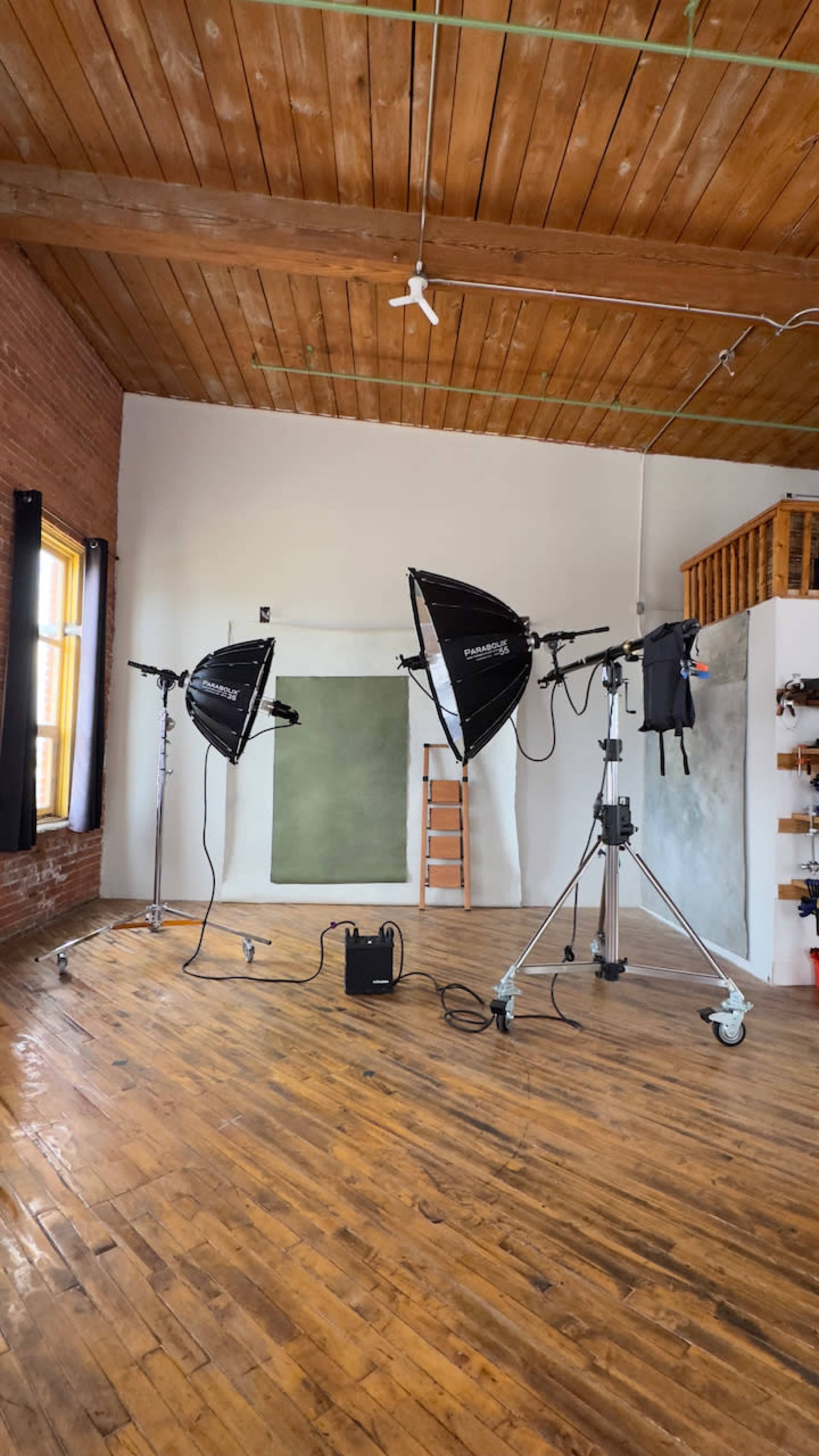 The image shows a photography studio featuring two light setups, a backdrop, a ladder, and a wooden floor.