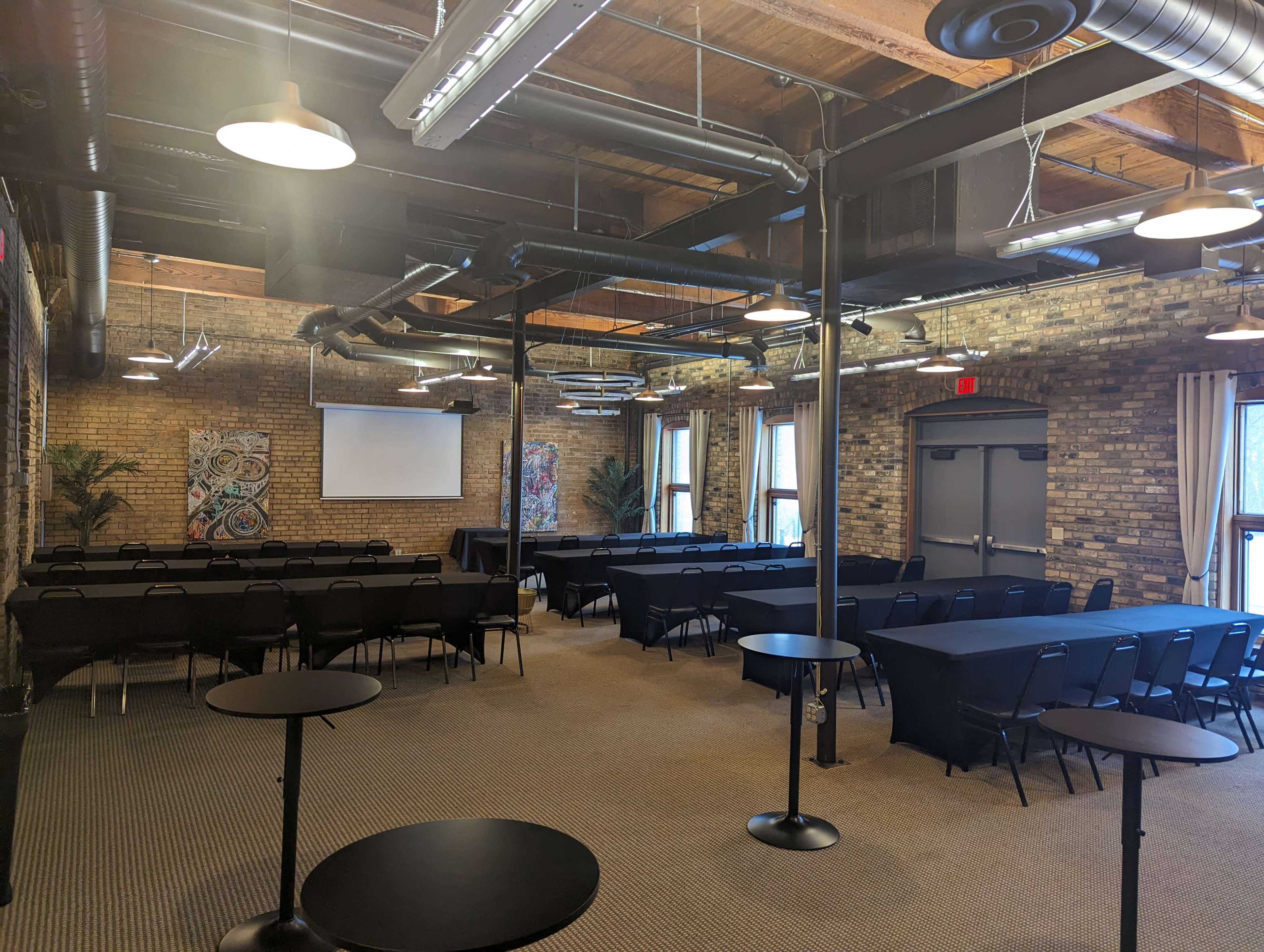 A spacious conference room features multiple rows of black tables and chairs, with a screen at the front and large windows allowing natural light.