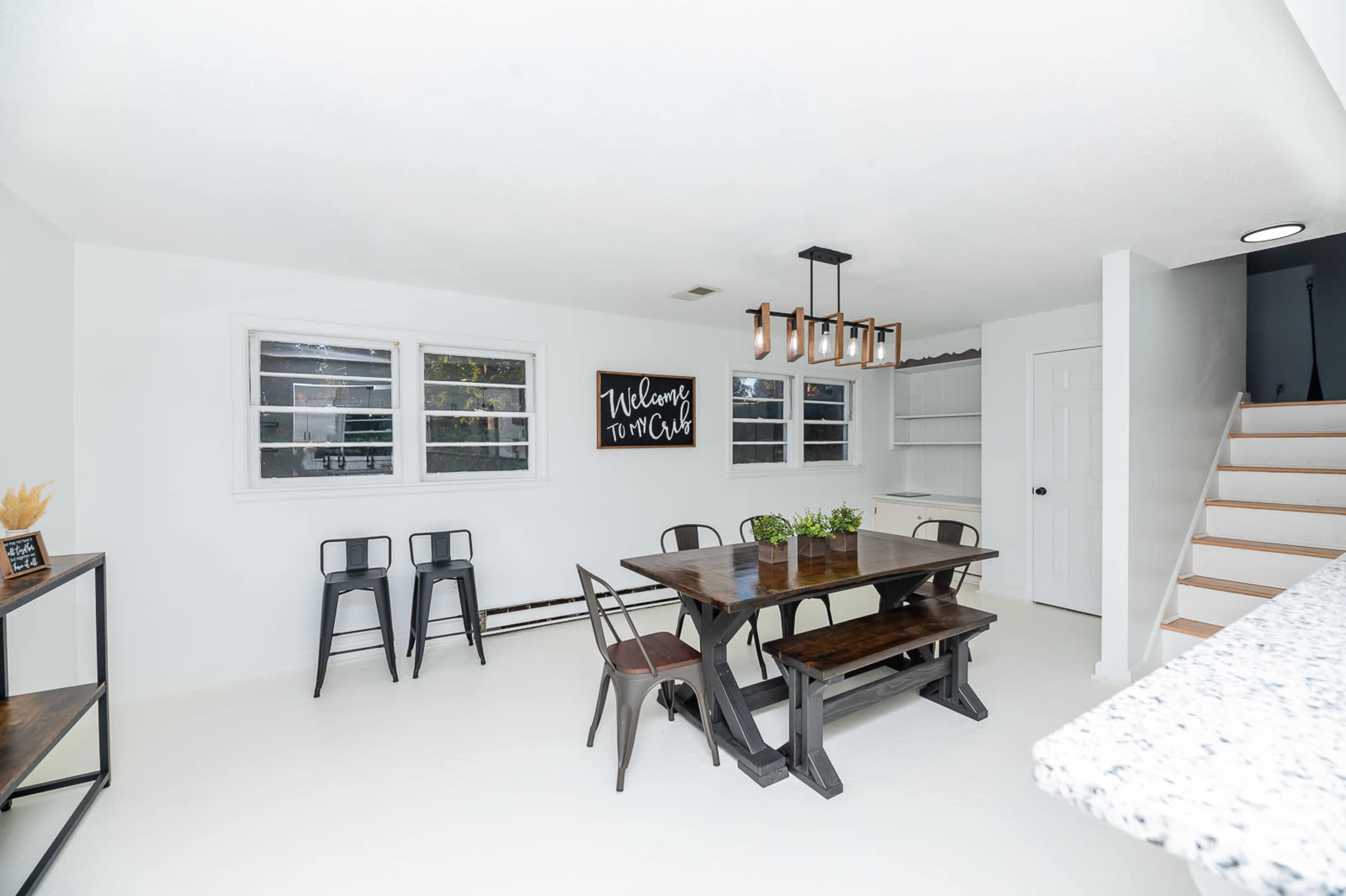 A contemporary dining area features a large wooden table surrounded by chairs, a wall-mounted sign, and a staircase leading to a second floor.