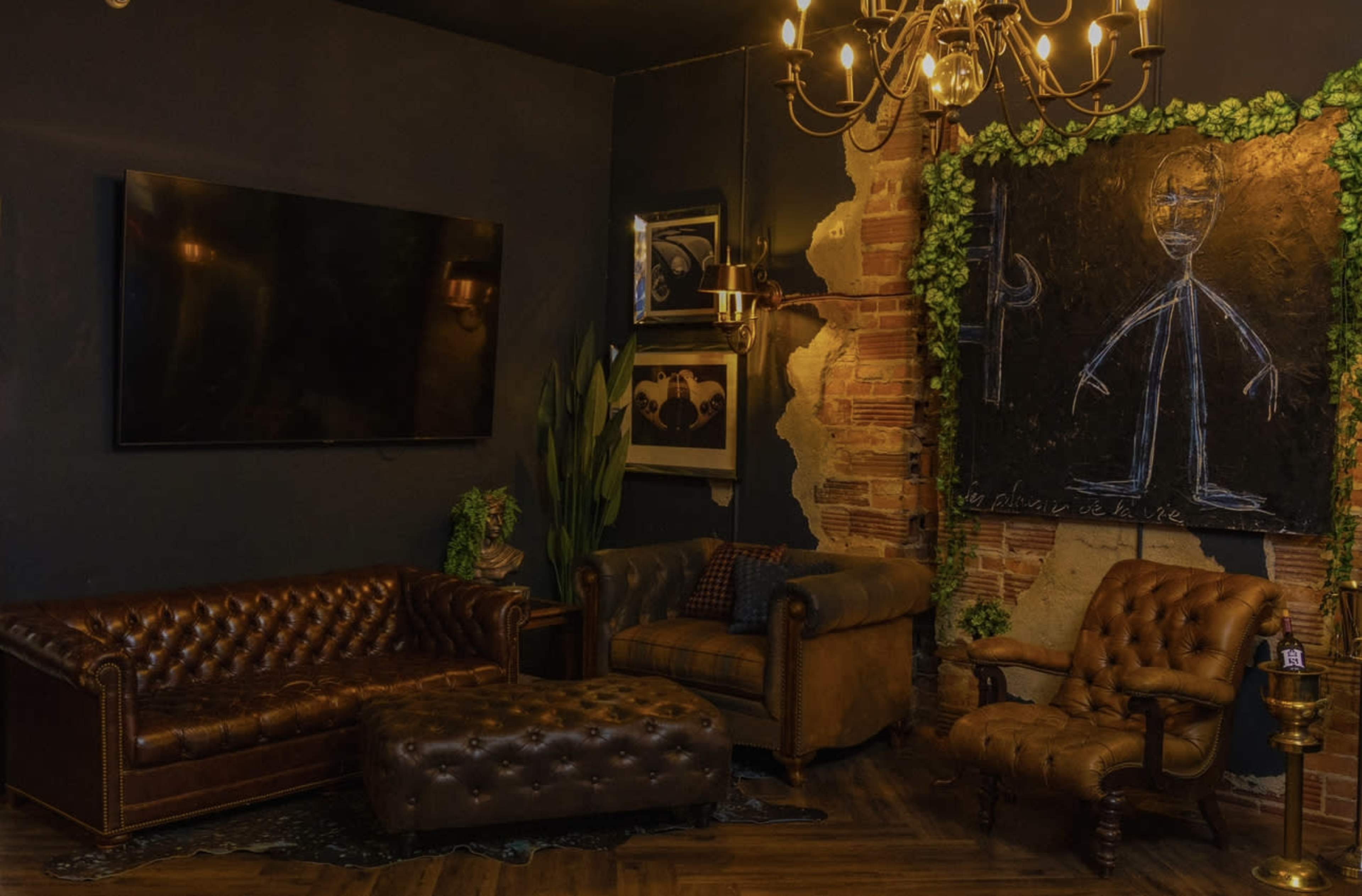 A cozy lounge area featuring a dark wall, a large flat-screen TV, a leather chesterfield sofa, an armchair, and a decorative blackboard surrounded by greenery.