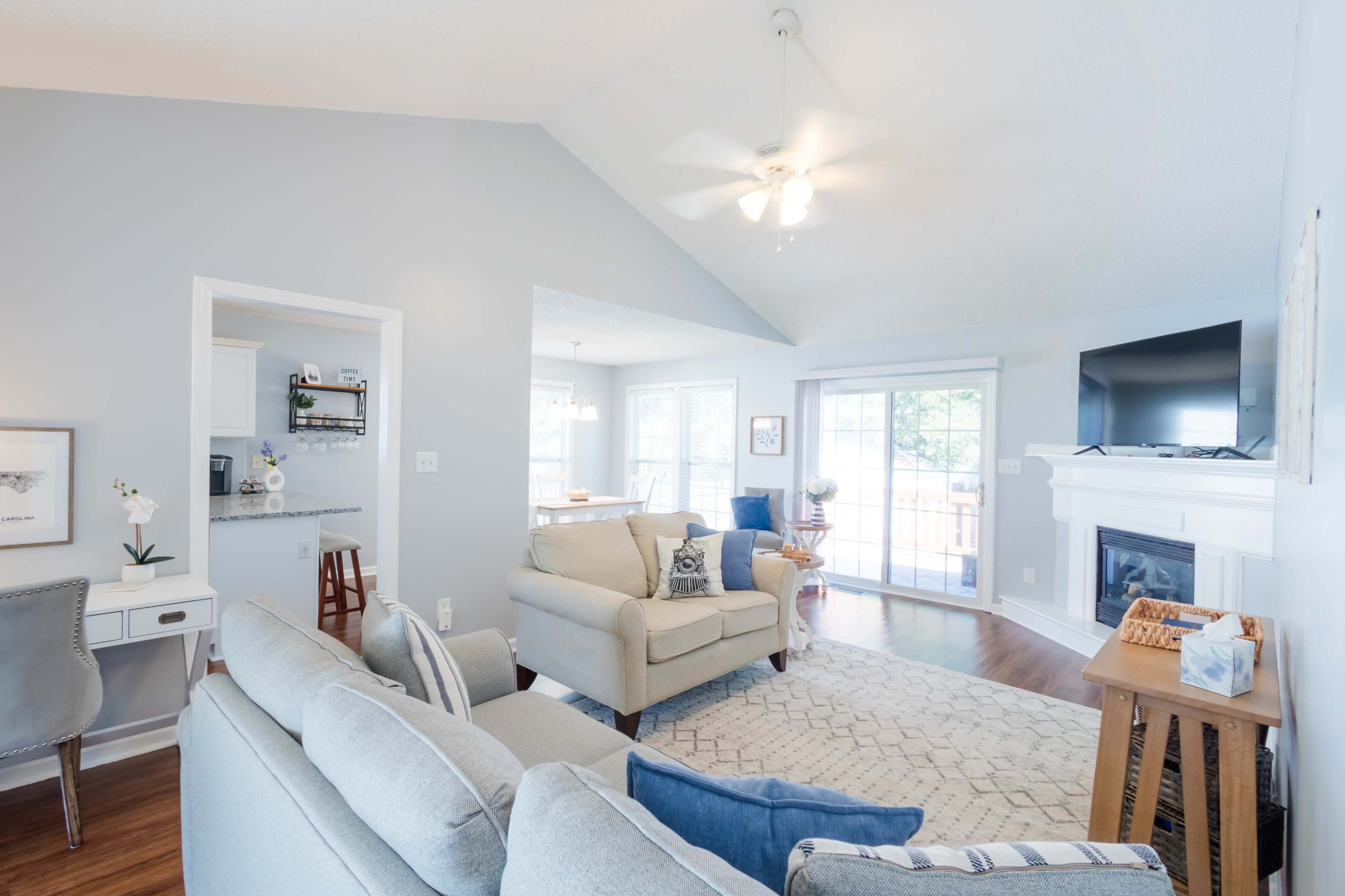 The image shows a bright living room with a light gray sofa, a coffee table, and large windows leading to a deck.