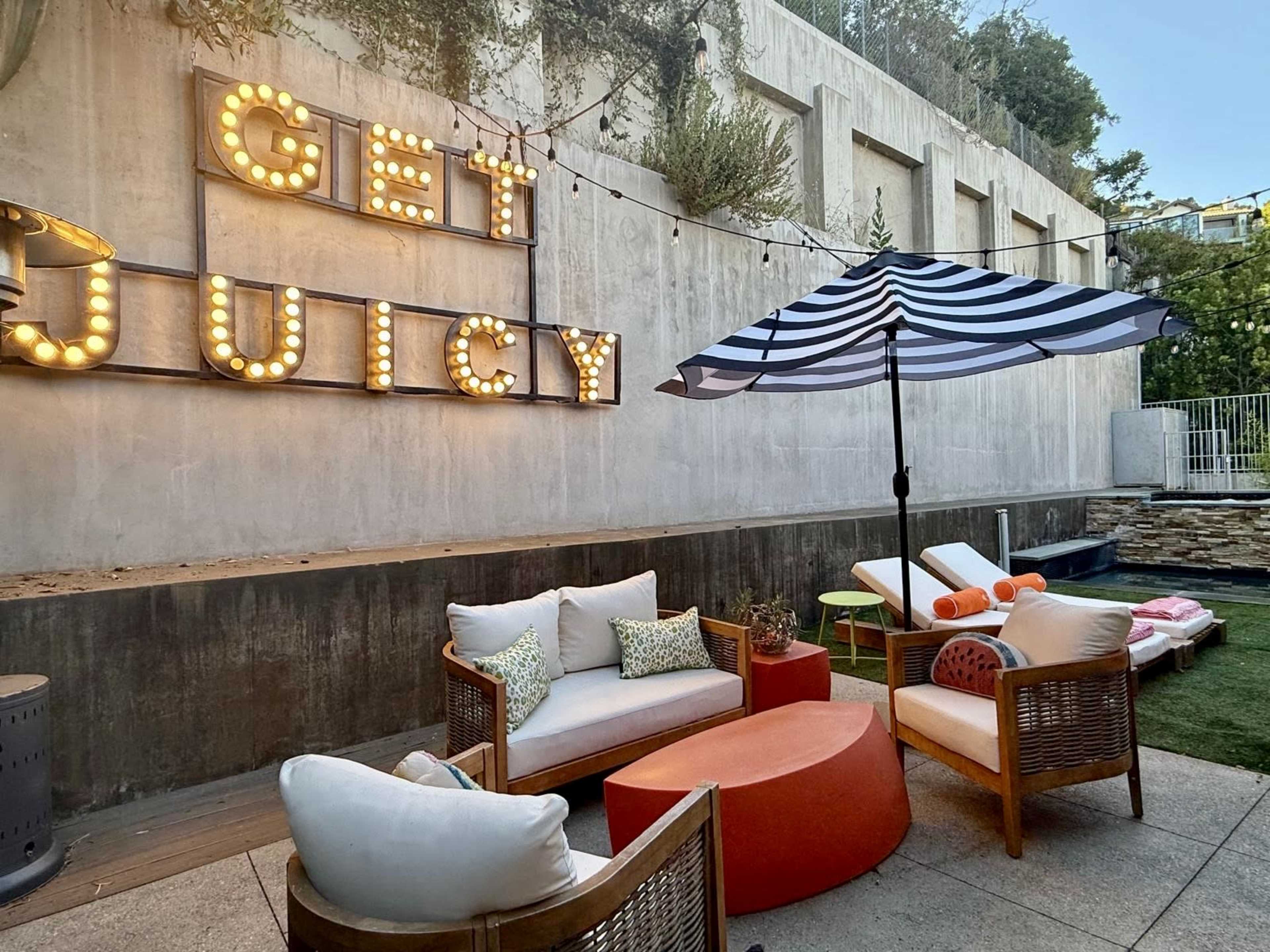 The image shows an outdoor lounge area featuring illuminated letters that spell "GET JUICY," a striped umbrella, and comfortable seating arrangements.
