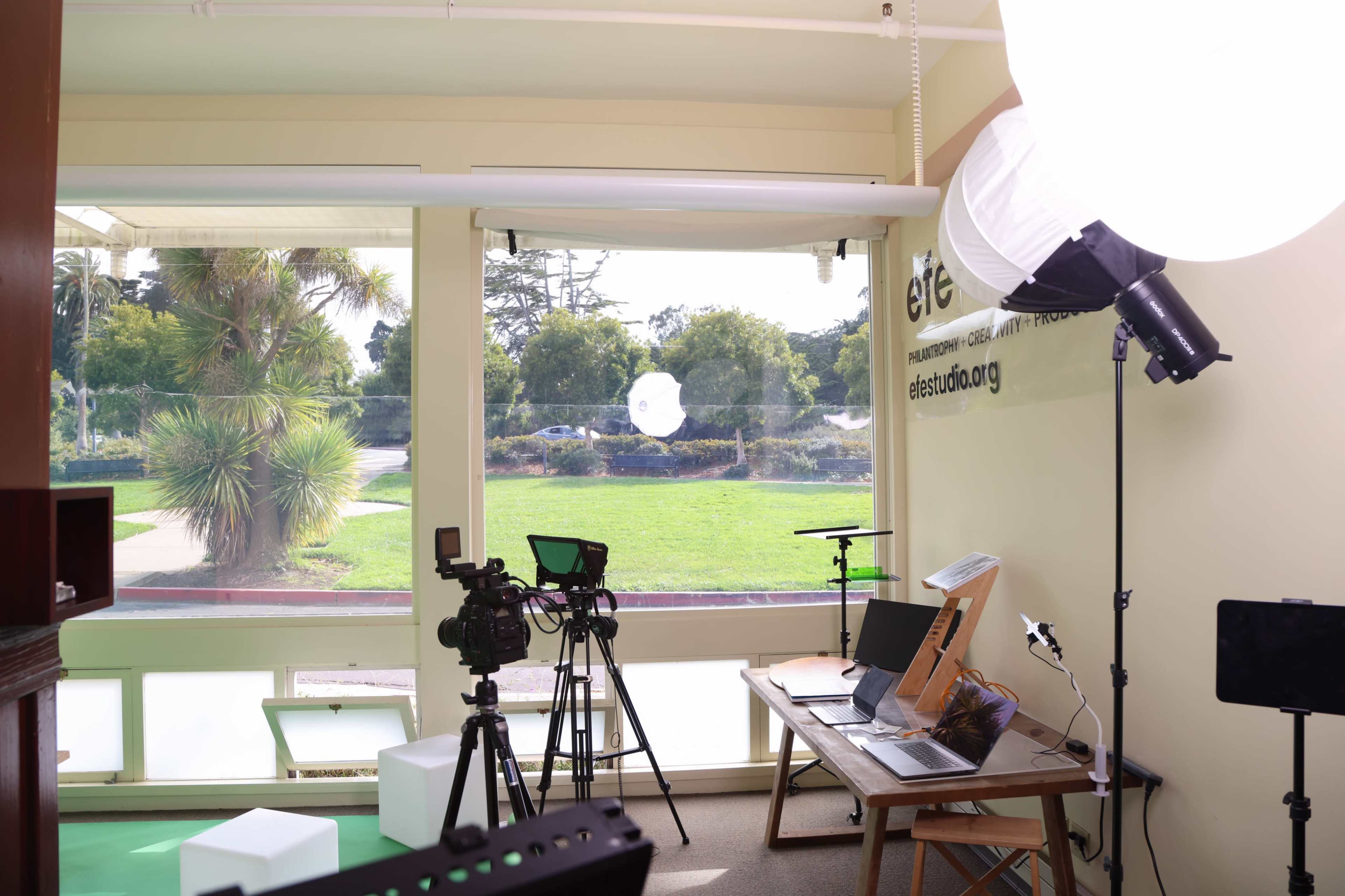 The image shows a bright room with a large window overlooking a green lawn, containing cameras, lighting equipment, and desks with laptops.