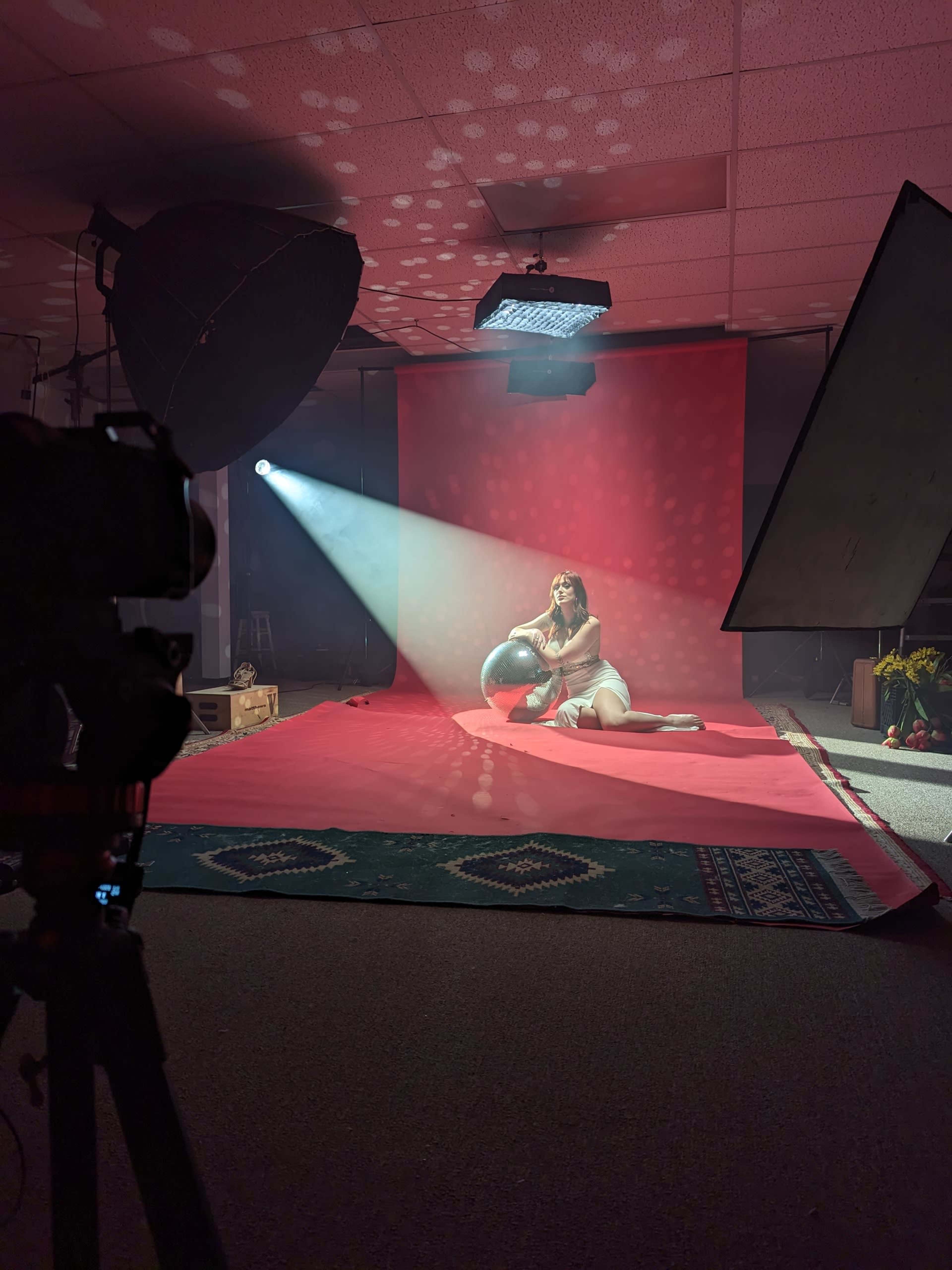 A model sits on a red backdrop in a photography studio, holding a globe while light beams illuminate the scene.